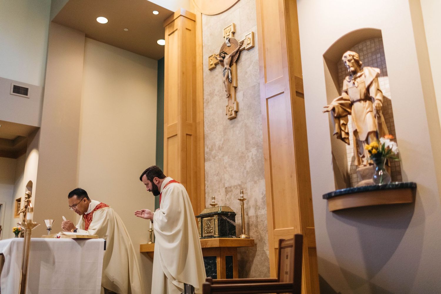 Catholic priests perform religious service at an altar with religious statues and decorative wall elements in background.
