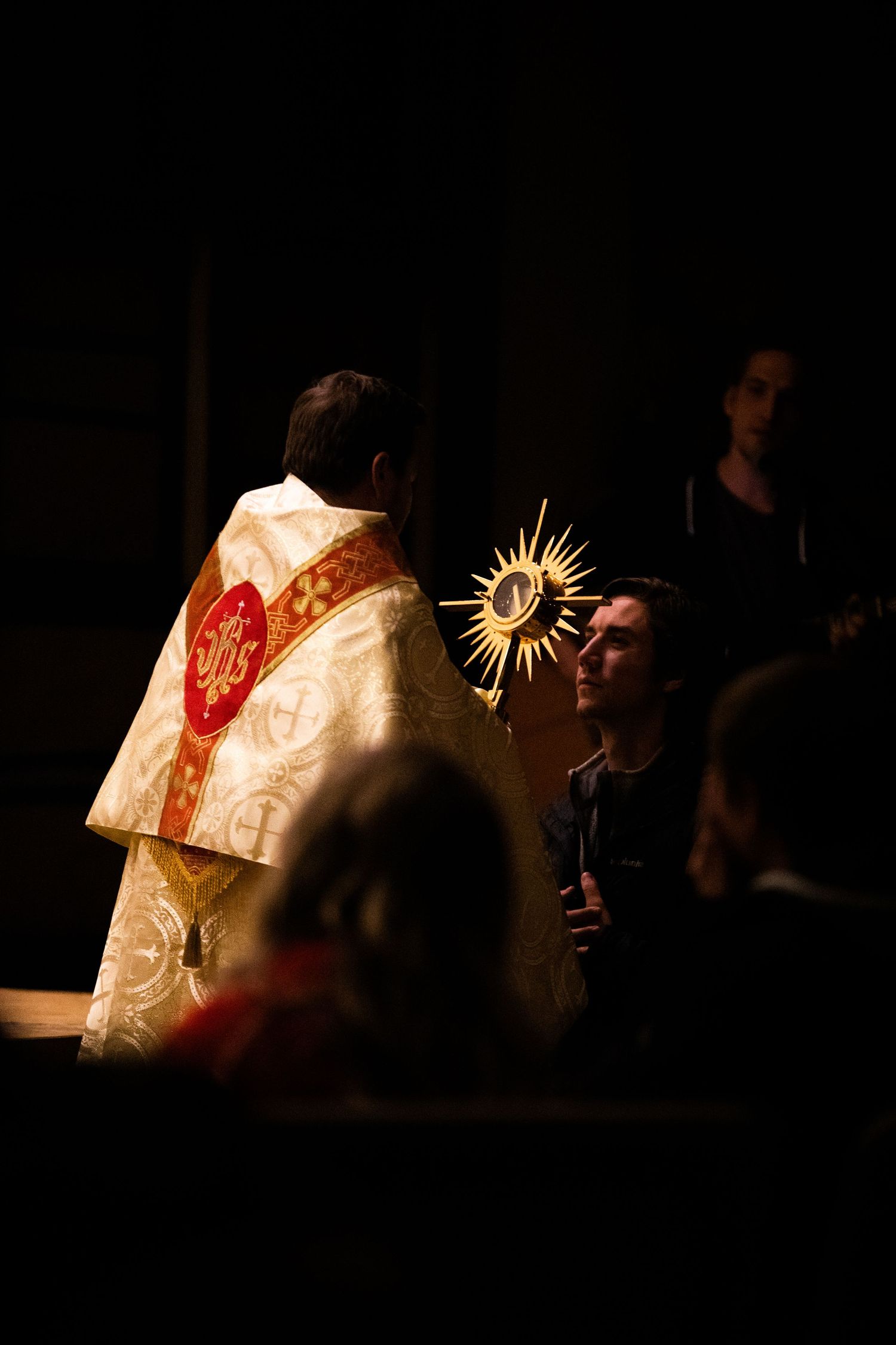 Catholic priest holding a golden monstrance during Eucharistic adoration in dramatic lighting.