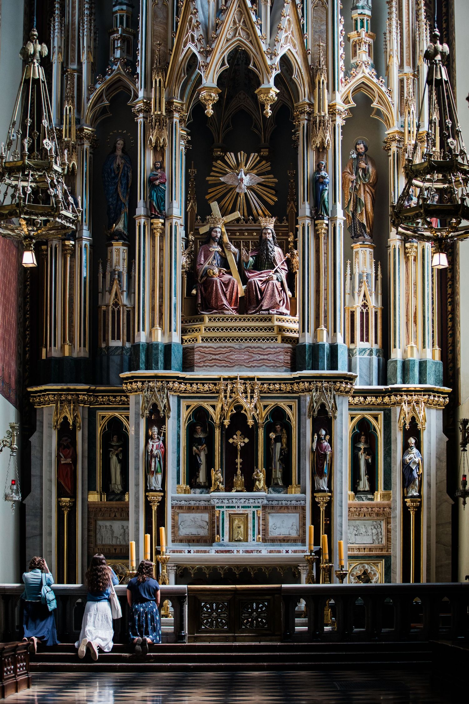 Worshippers kneeling before an elaborate Gothic church altar adorned with religious statues and gold details.