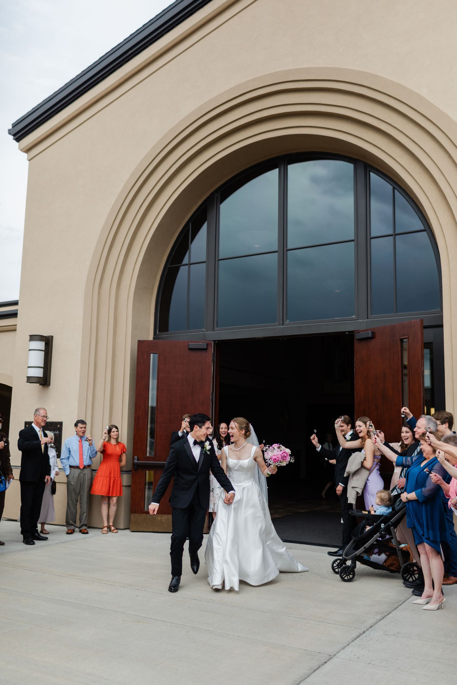 Wedding party exits through arched doorway of modern building as guests gather outside to celebrate.