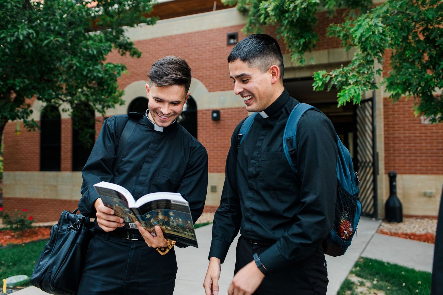 Students in dark suits socializing outdoors on a college campus with brick buildings and trees visible.