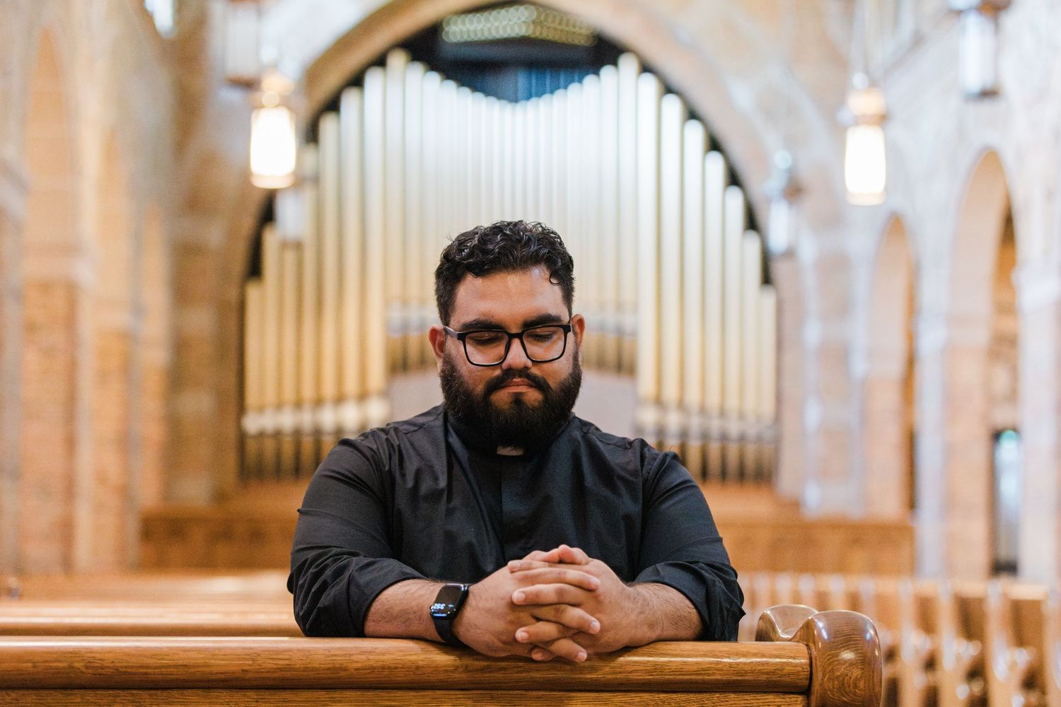 Bearded man in black shirt sits in church pew with large pipe organ in background.