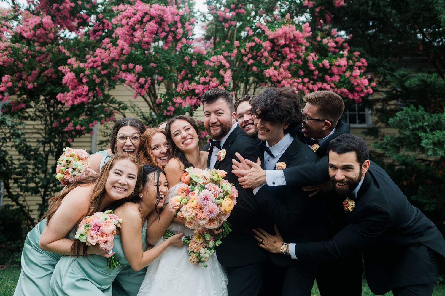 Joyful wedding party laughing together outdoors with pink flowering trees, bride holding colorful bouquet in white gown.