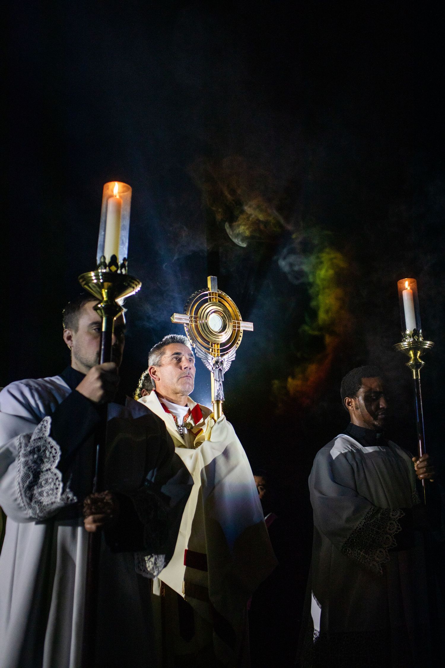 Religious procession in white robes carrying candles and liturgical items in a dark setting with dramatic lighting.