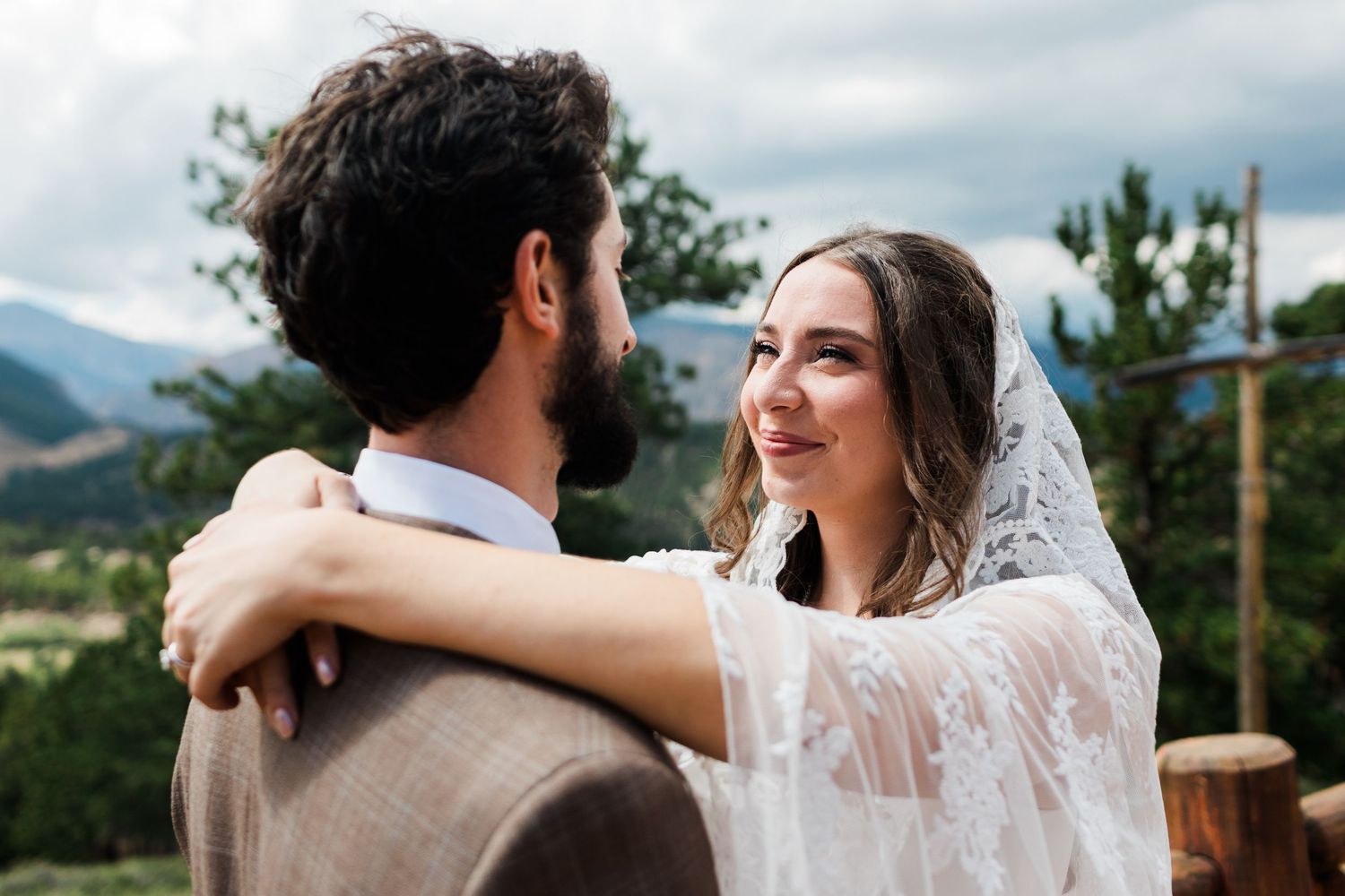 Bride in lace veil embraces groom outdoors with mountain views, sharing a romantic wedding moment.