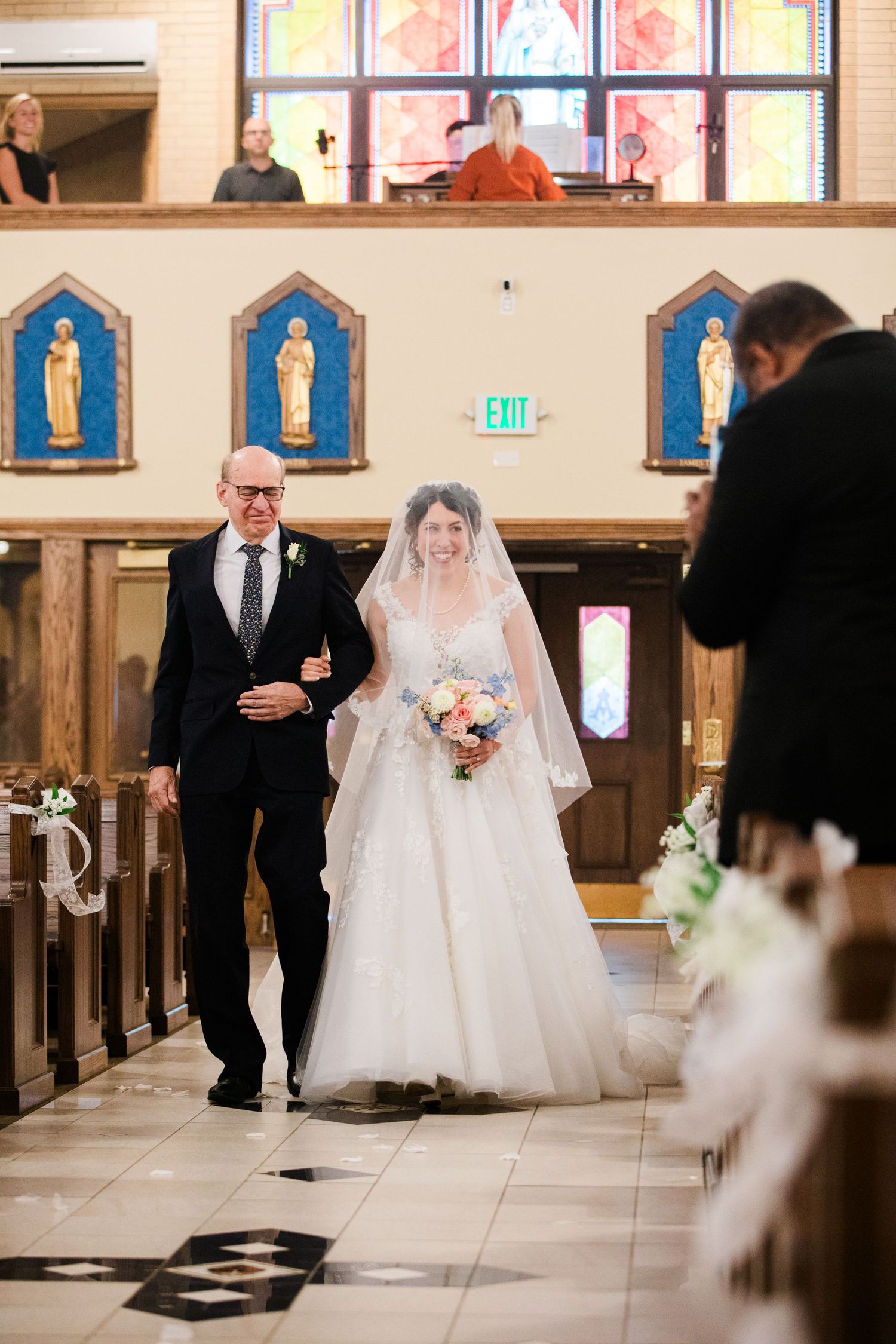 Bride walks down church aisle with escort, wearing white gown and veil, holding bouquet, passing wooden pews and stained glass.