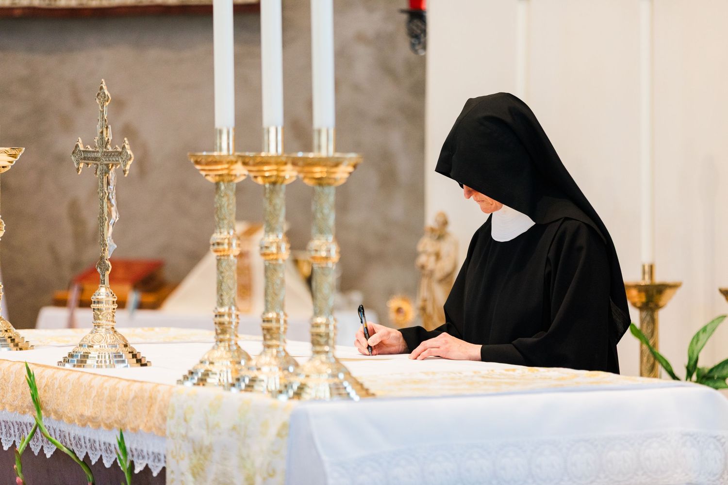 A nun in black habit signs documents at an altar adorned with golden candelabras during a religious ceremony.