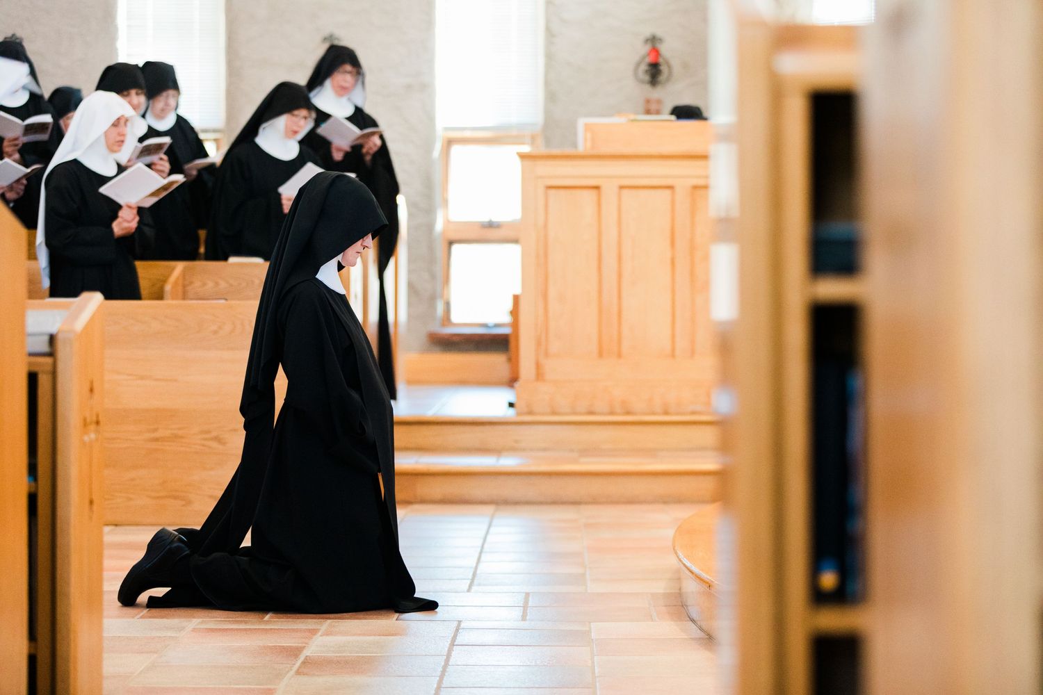 Catholic nuns in traditional black habits kneeling and praying in a modern wooden church chapel.