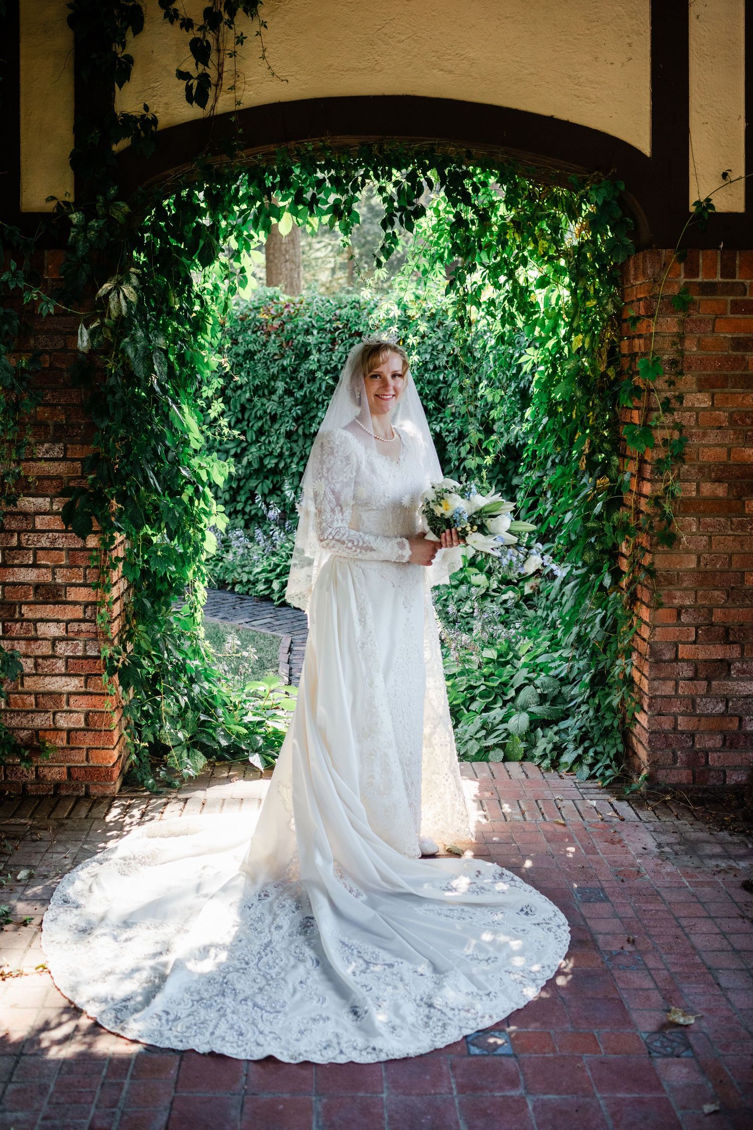 Bride in lace wedding gown with veil holds bouquet under ivy-covered brick arch in garden.