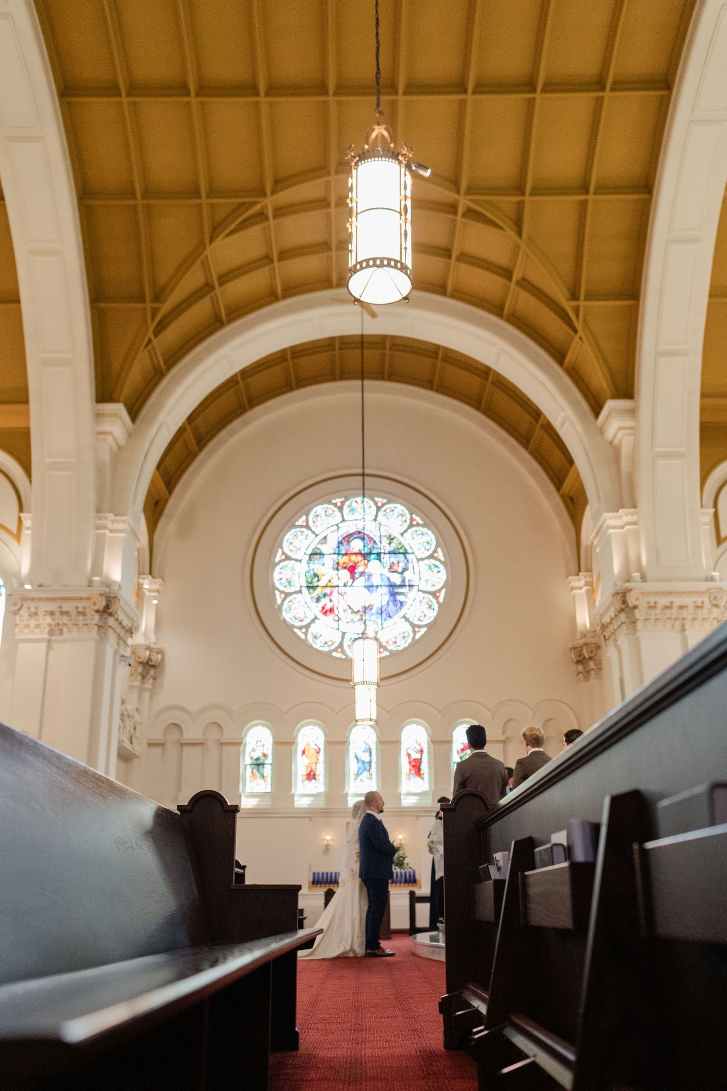 Couple exchanging vows at church altar with stunning stained glass rose window and vaulted wooden ceiling.