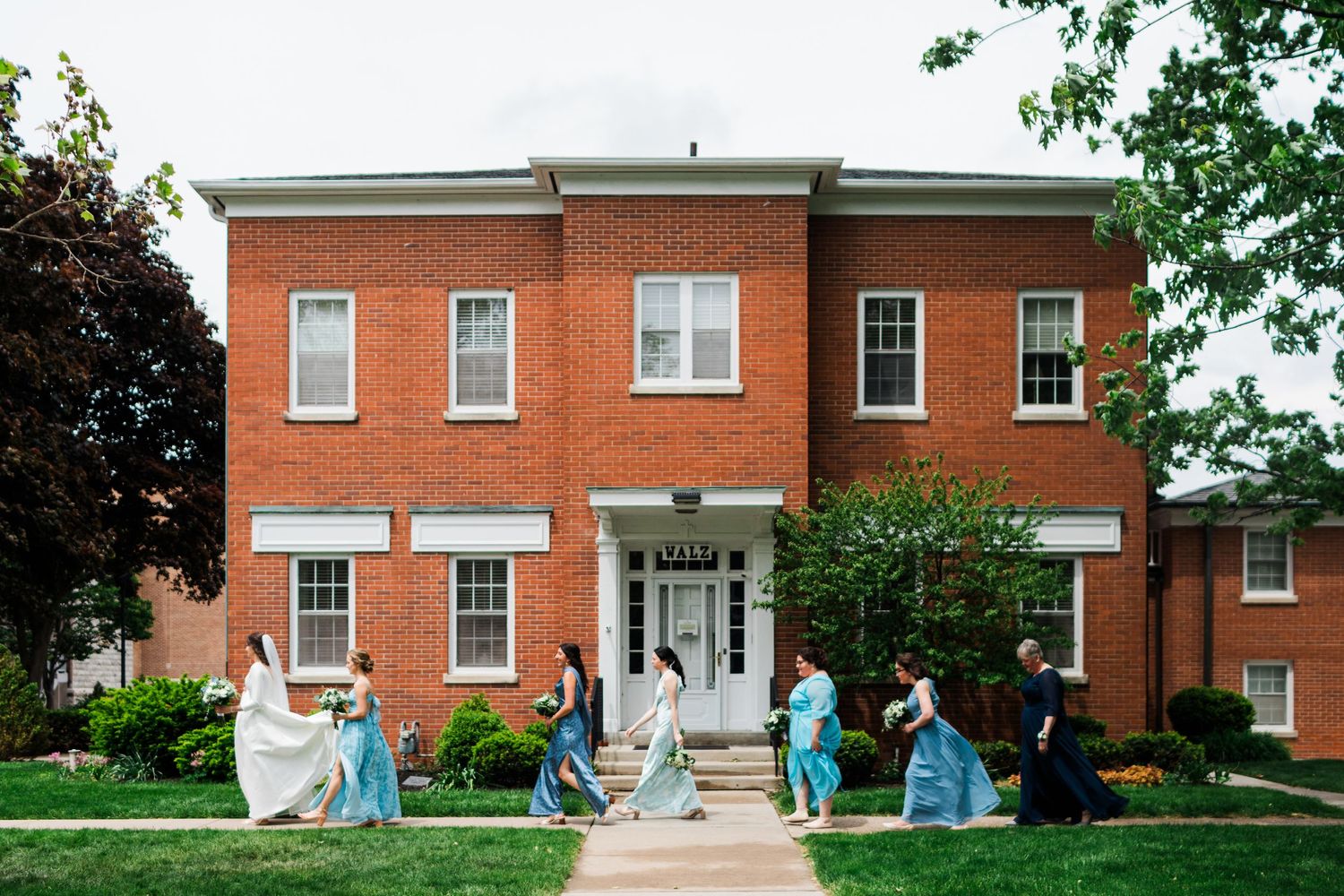 Wedding party walking in front of a red brick building, bridesmaids in blue gowns with bride in white dress.
