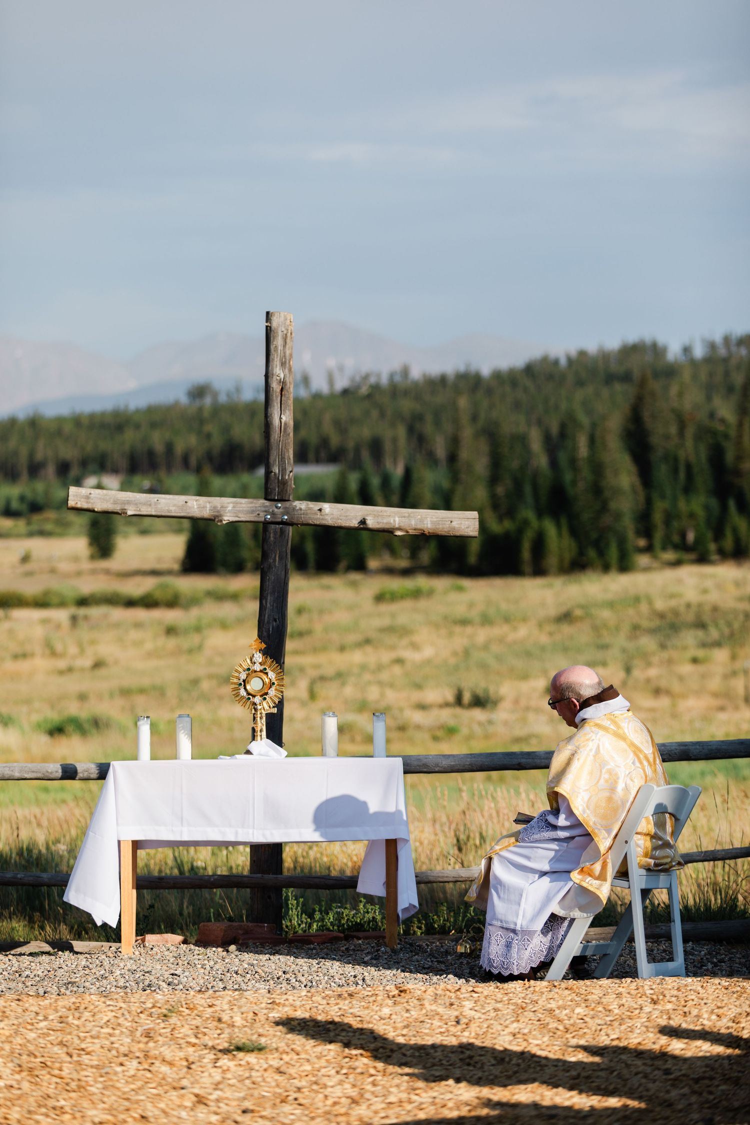 A wooden cross memorial stands in a grassy field with a forest backdrop and wood chips scattered on the ground.