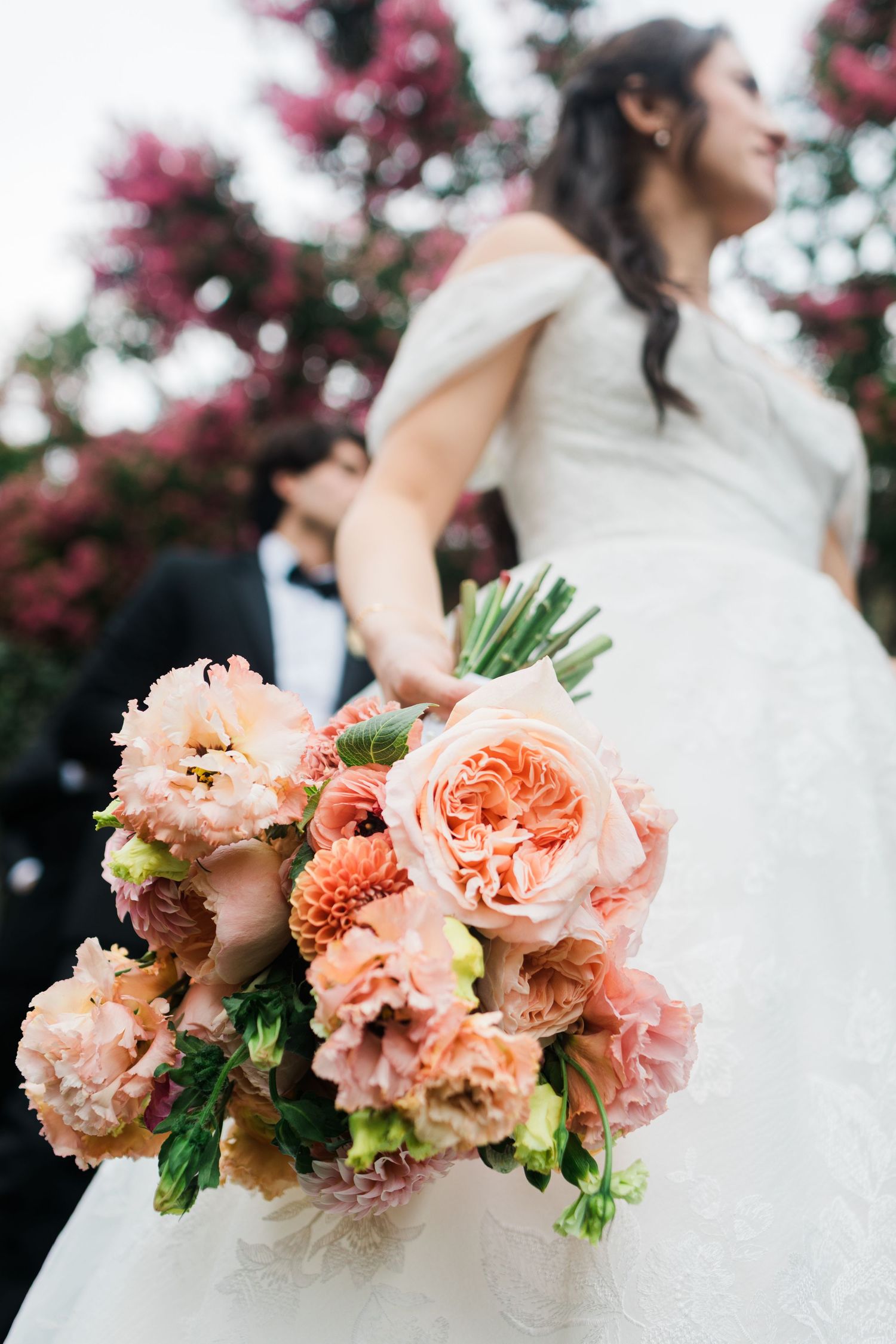 Bride holding a lush pink and peach bouquet with roses and dahlias, groom in tuxedo behind her near blooming trees.