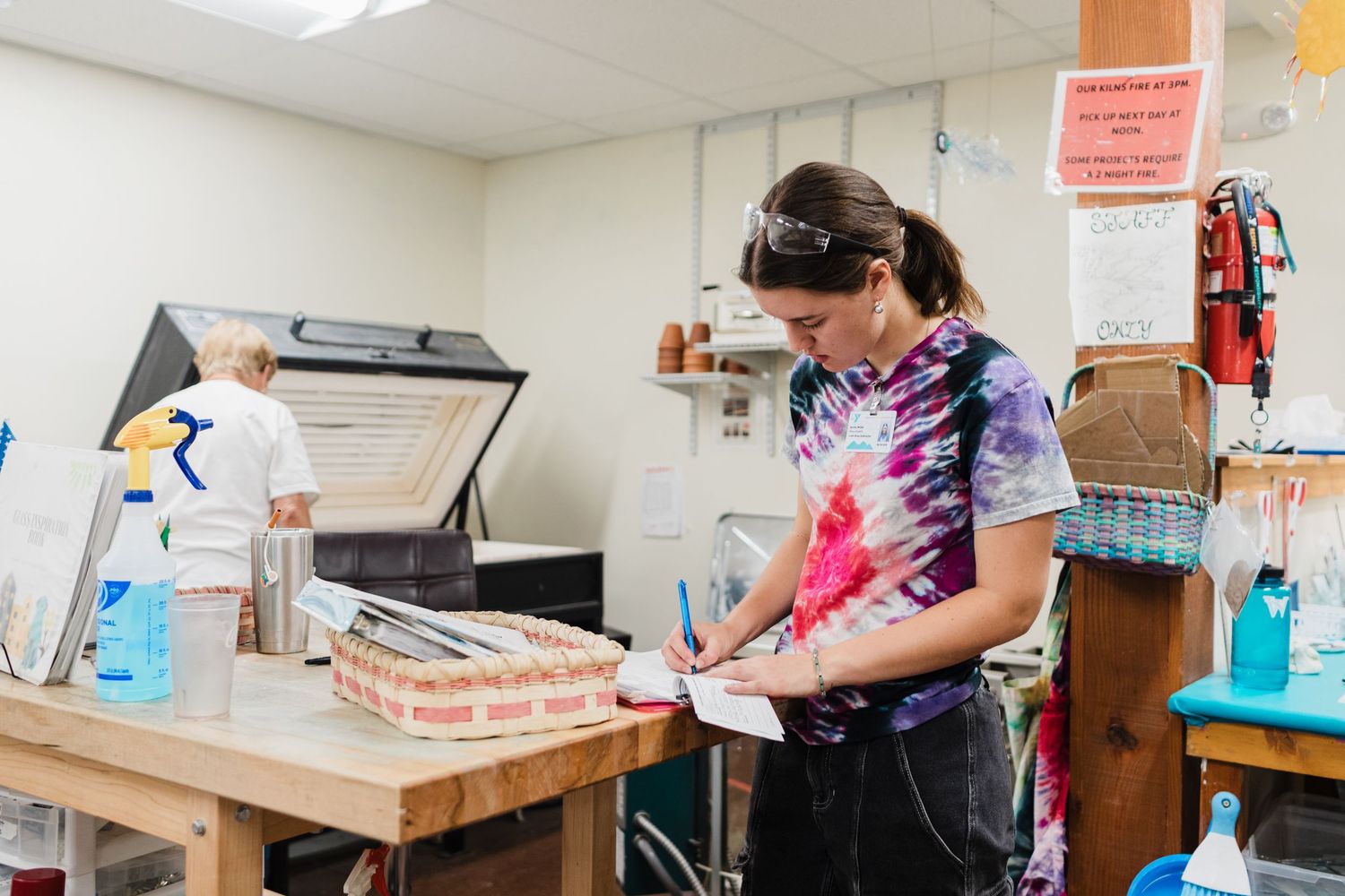 Woman in tie-dye shirt works at a craft table in an art studio with printing equipment in the background.