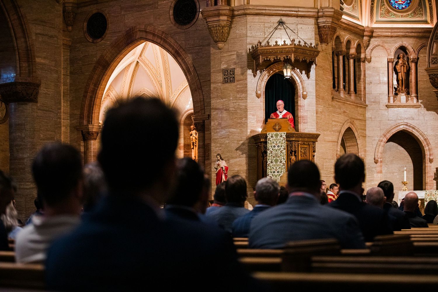 Congregation sits in wooden pews listening to a speaker at a podium inside a grand stone cathedral.