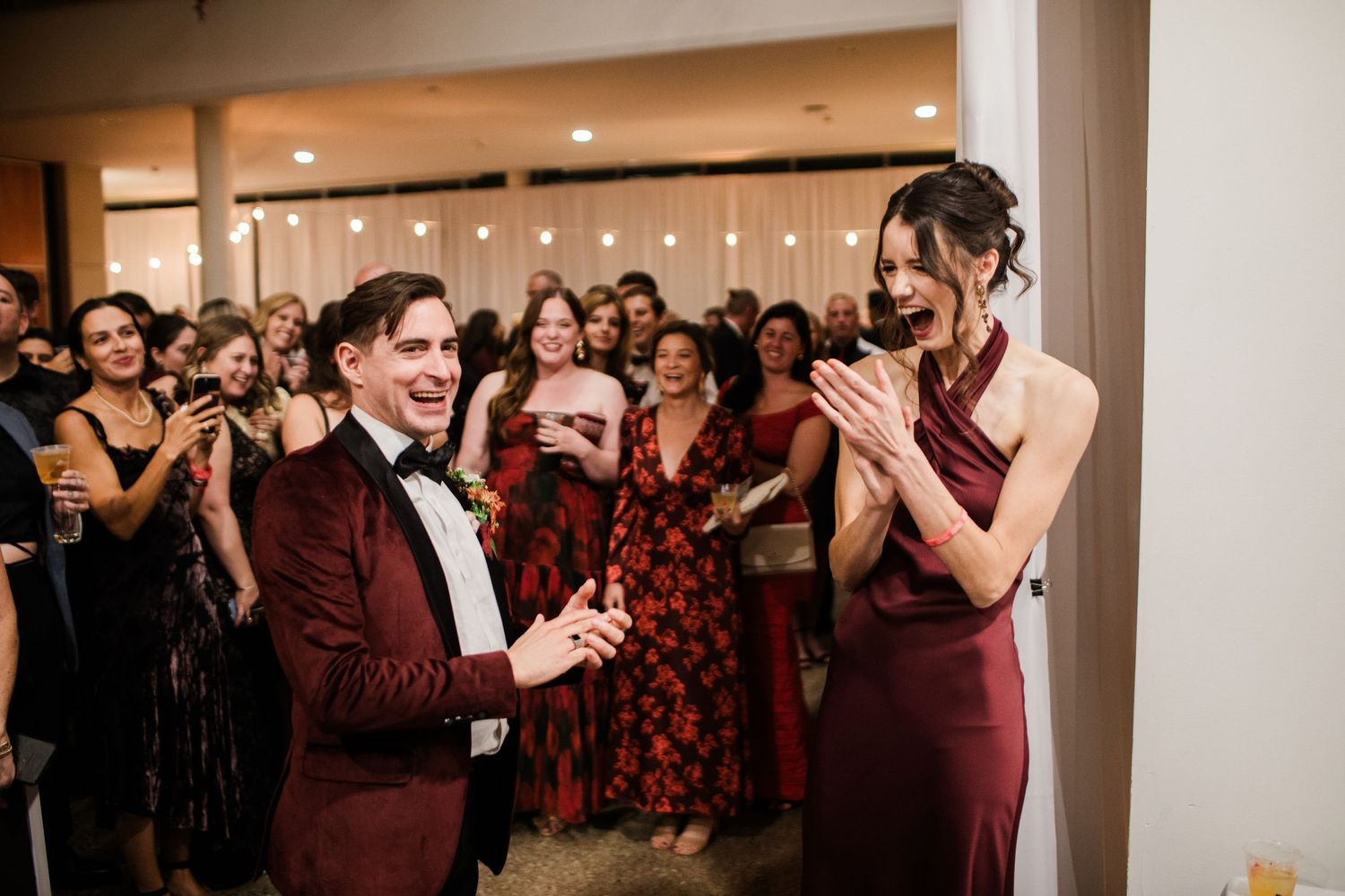 Two people in burgundy formal attire share a special moment during an event surrounded by guests.