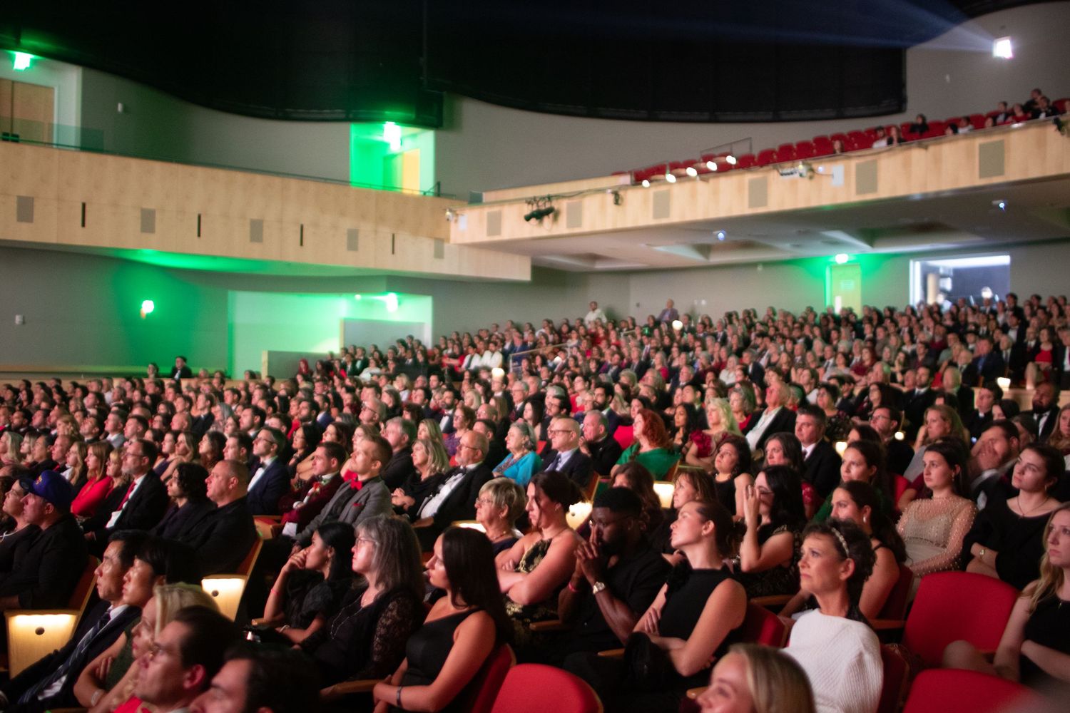 A packed theater auditorium filled with audience members watching a performance in red seats with green lighting on walls.