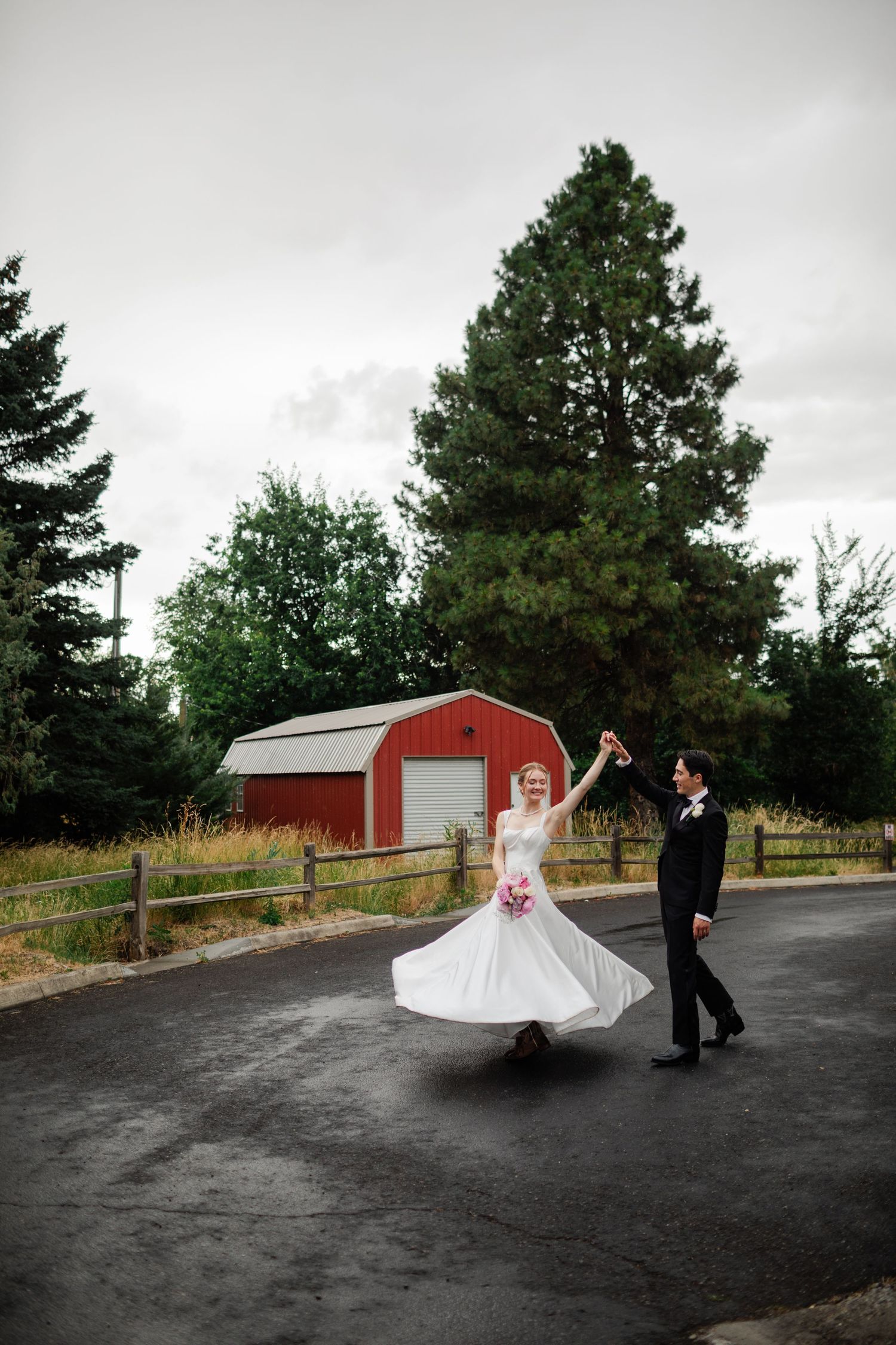 A wedding couple poses for creative photos on a black driveway with a red barn in the background.