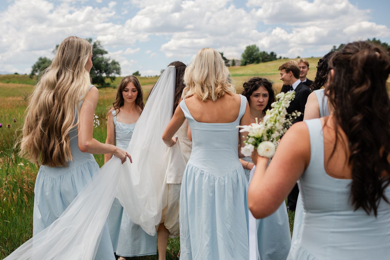 Bridesmaids in light blue dresses gather around the bride in a sunny outdoor field.