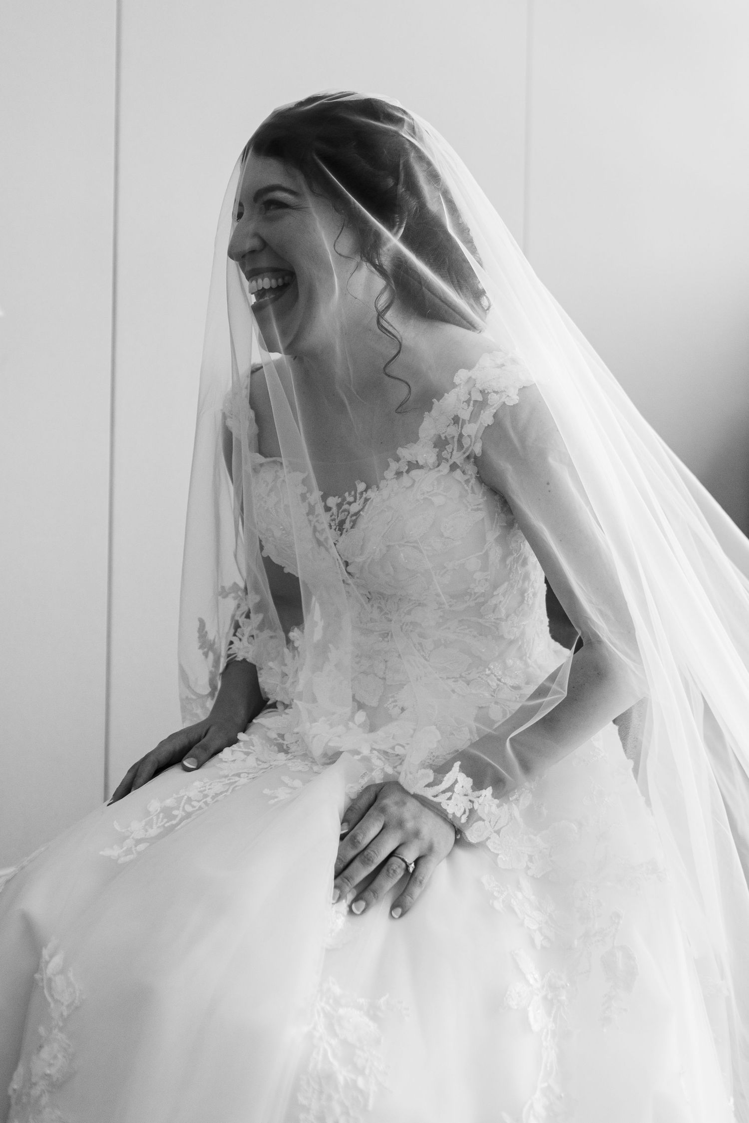 Black and white photo of a laughing bride in a lace wedding gown and veil, radiating joy on her wedding day.