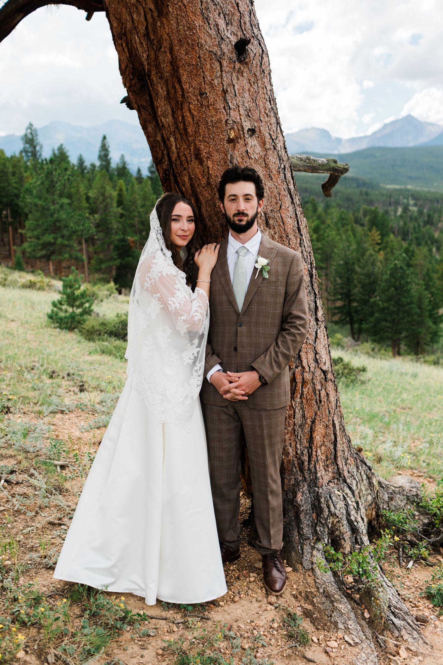 Bride and groom pose by a pine tree with mountain forest backdrop on their wedding day.