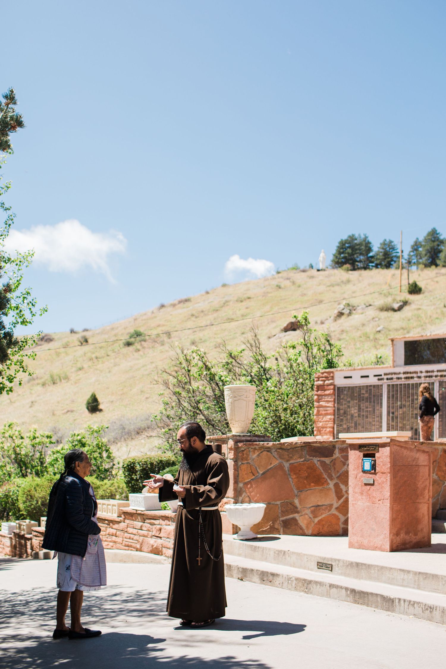 A Franciscan friar in brown robes speaks with a visitor outdoors at a sunny hillside religious shrine.