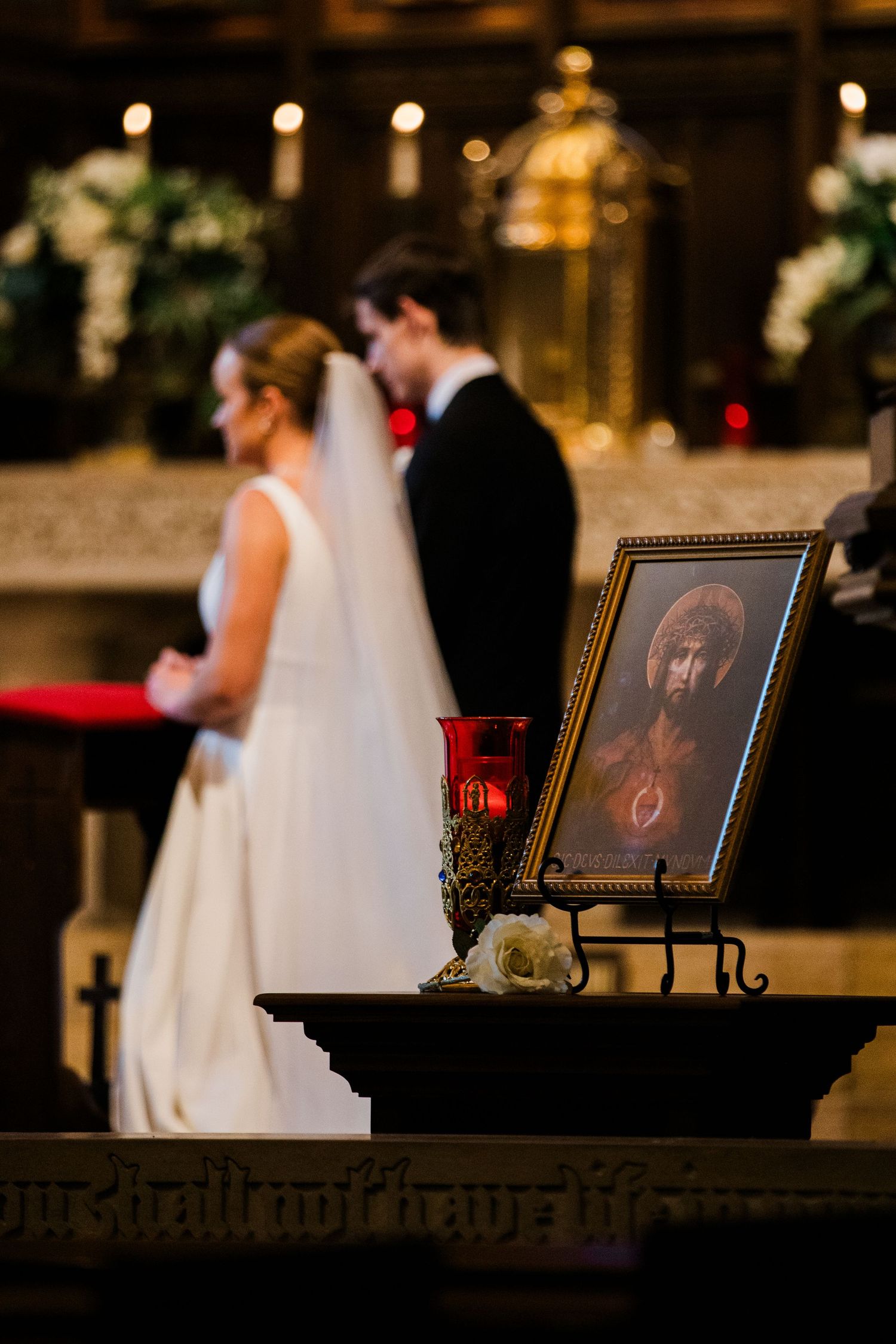 Bride and groom at Catholic church altar with sacred heart icon and red candle in foreground during wedding ceremony.
