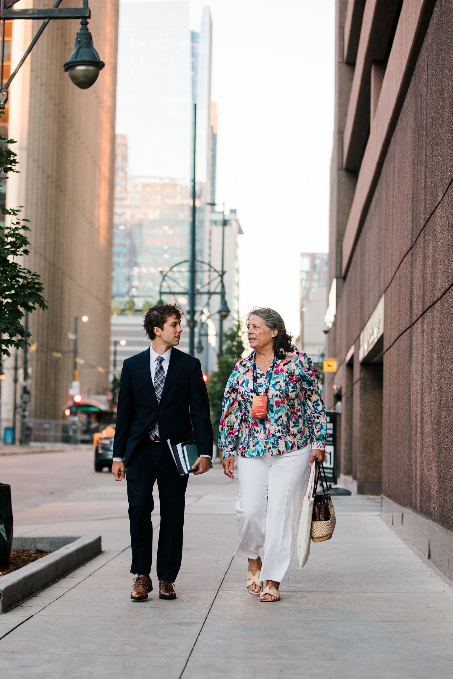 A young businessman and an older woman in floral jacket walk together on an urban city sidewalk.