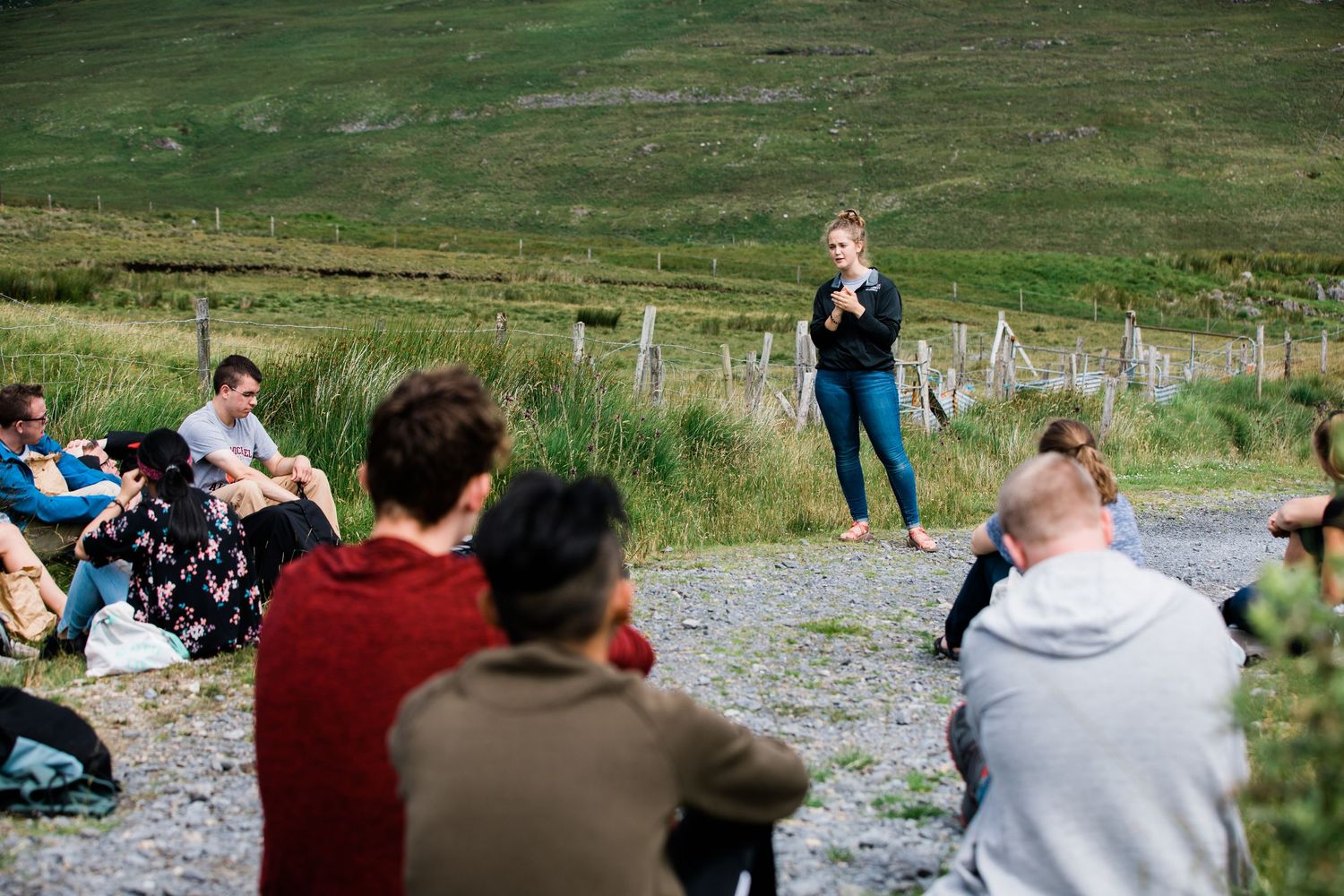 A young woman speaks to a group of students sitting outdoors in an Irish hillside setting.