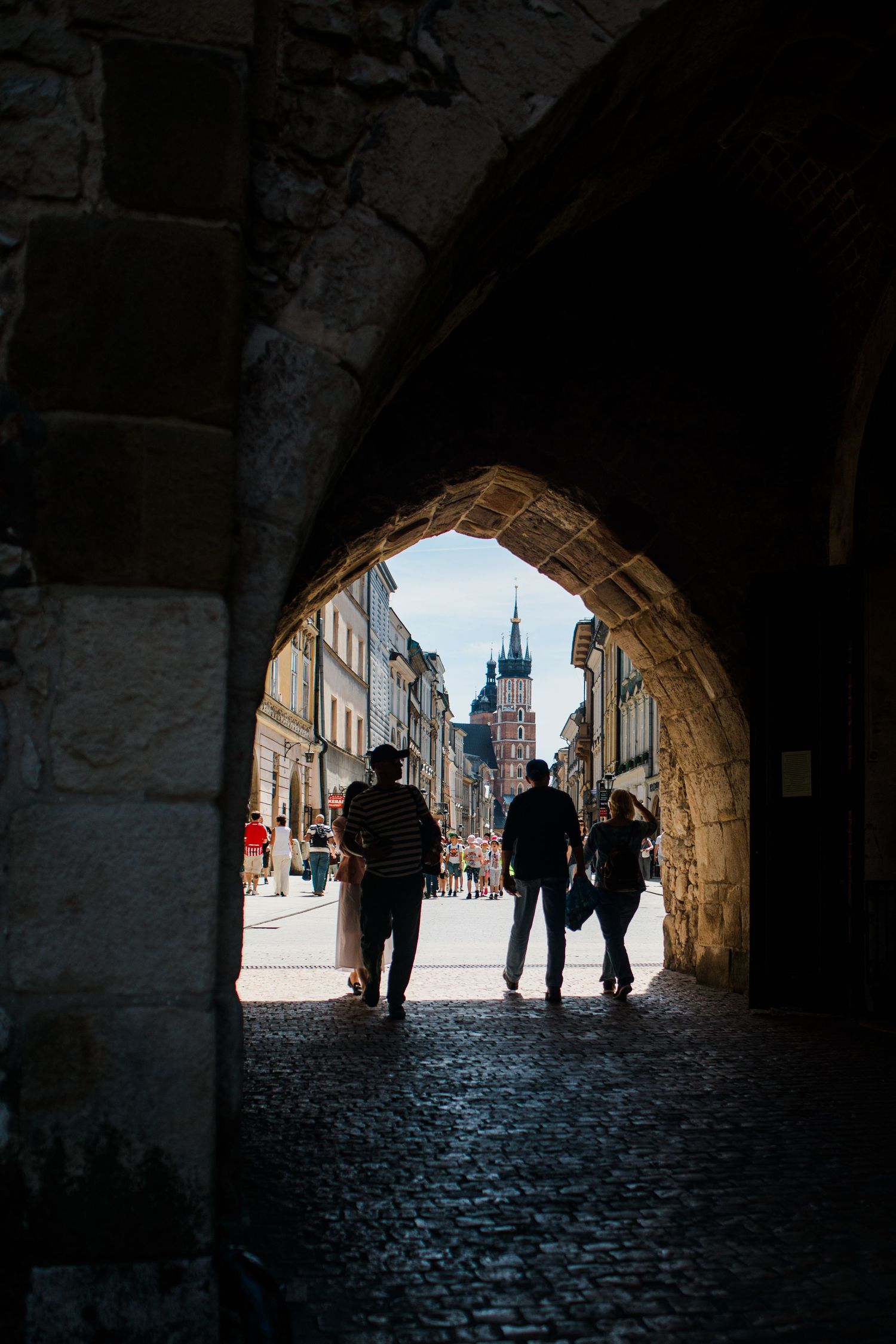Silhouetted tourists walking through medieval stone arch gateway toward St. Marys Basilica in Krakow.