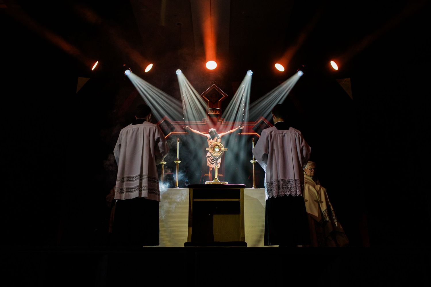 A dramatic stage performance features dancers in white costumes under dramatic spotlighting and theatrical fog effects.
