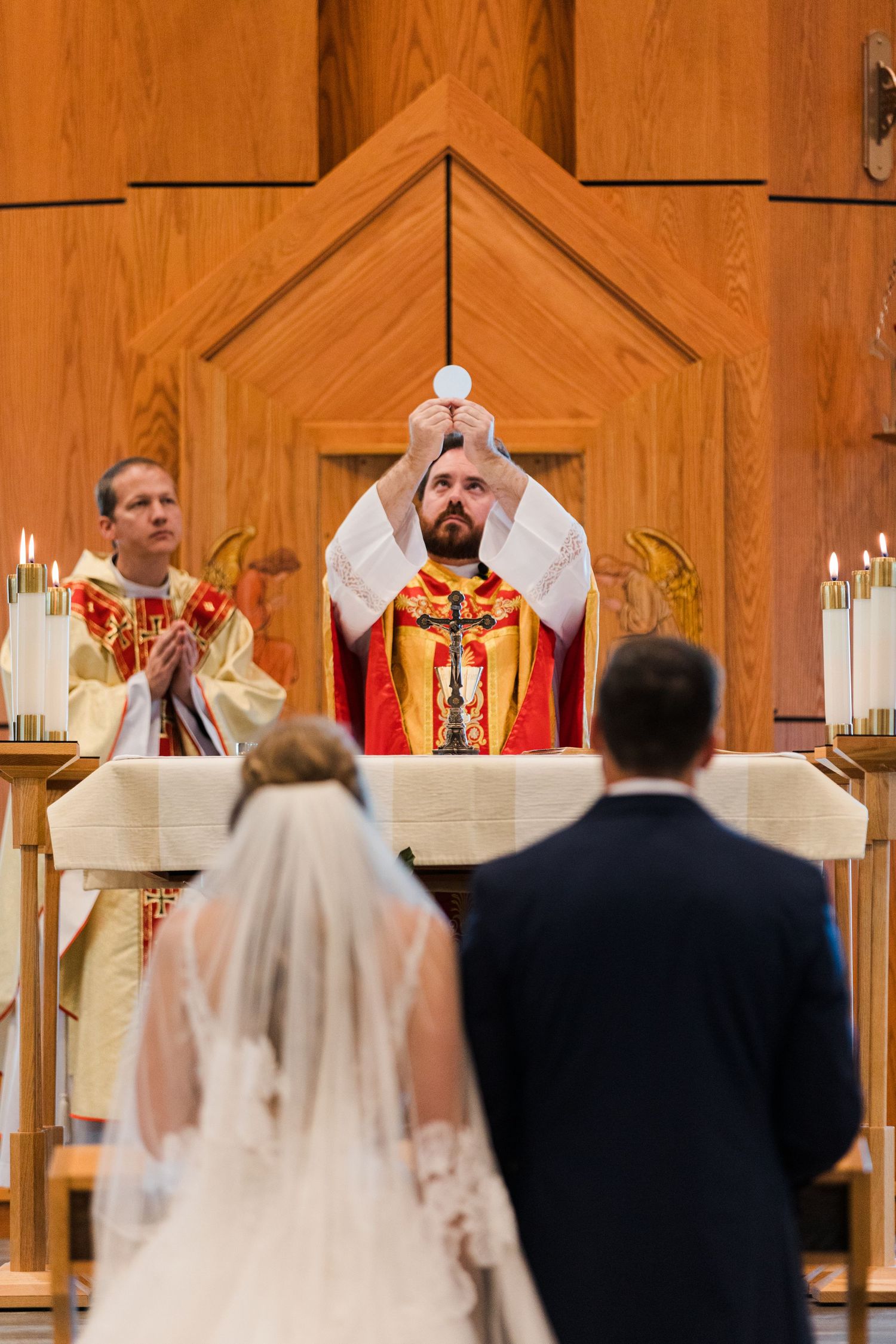 Catholic priest elevates the Eucharist during a wedding Mass ceremony with bride and groom standing before the altar.