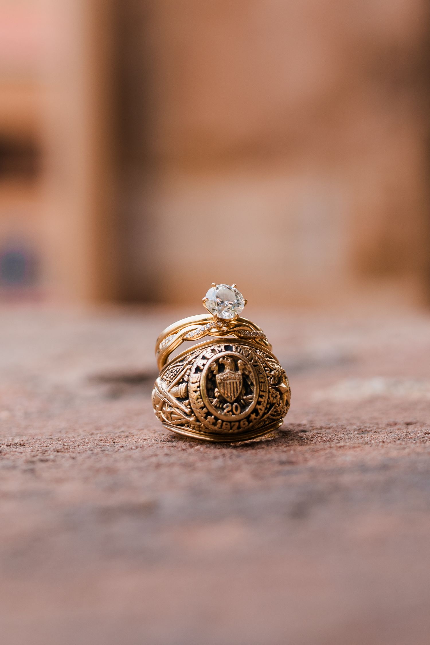 Gold class ring with diamond engagement ring stacked on rustic brick surface, wedding jewelry detail.