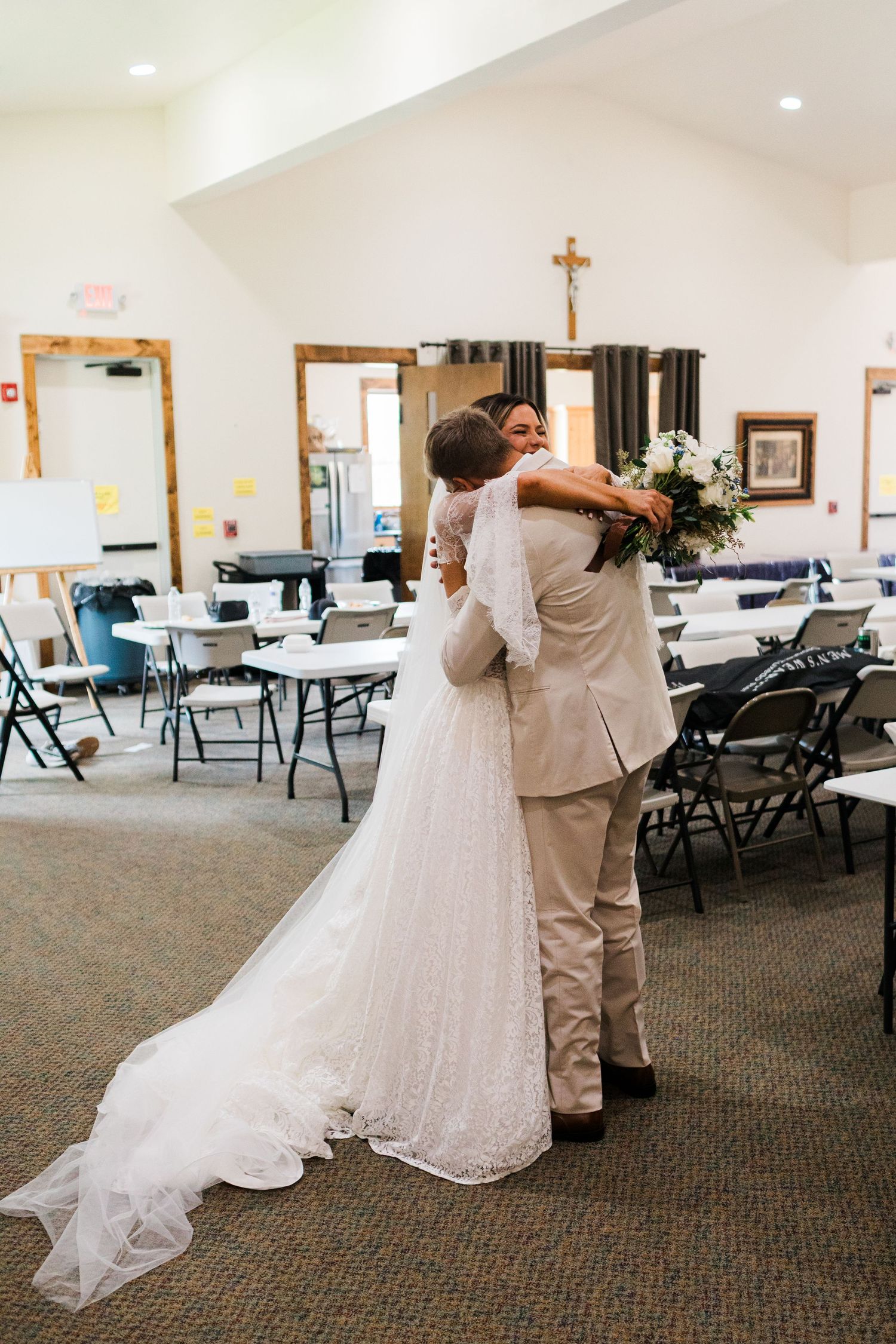 Bride and groom share a heartfelt hug in a church hall, bride holding a bouquet in a lace gown with a long veil.