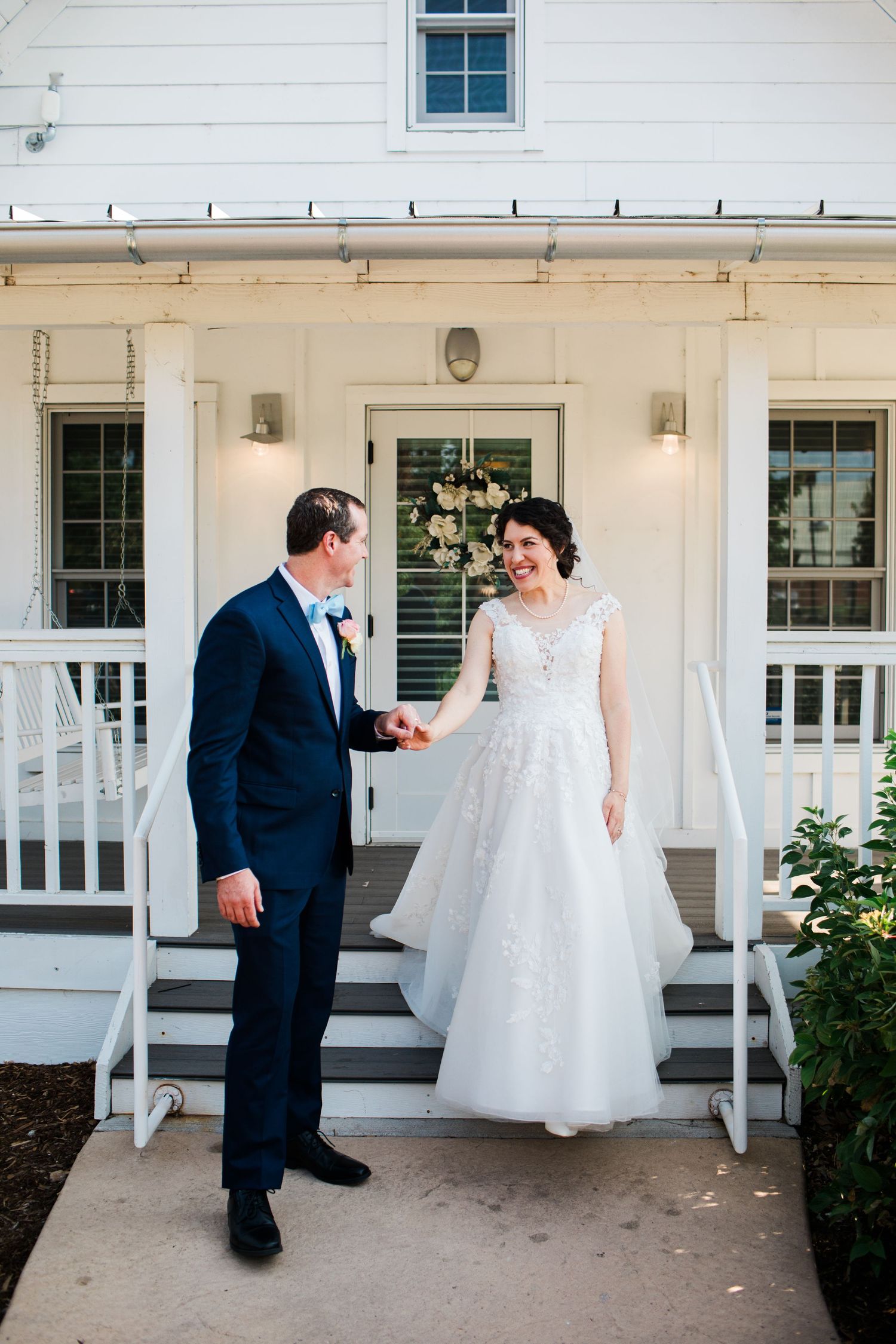 A bride and groom hold hands and smile at each other in front of a white farmhouse on their wedding day.