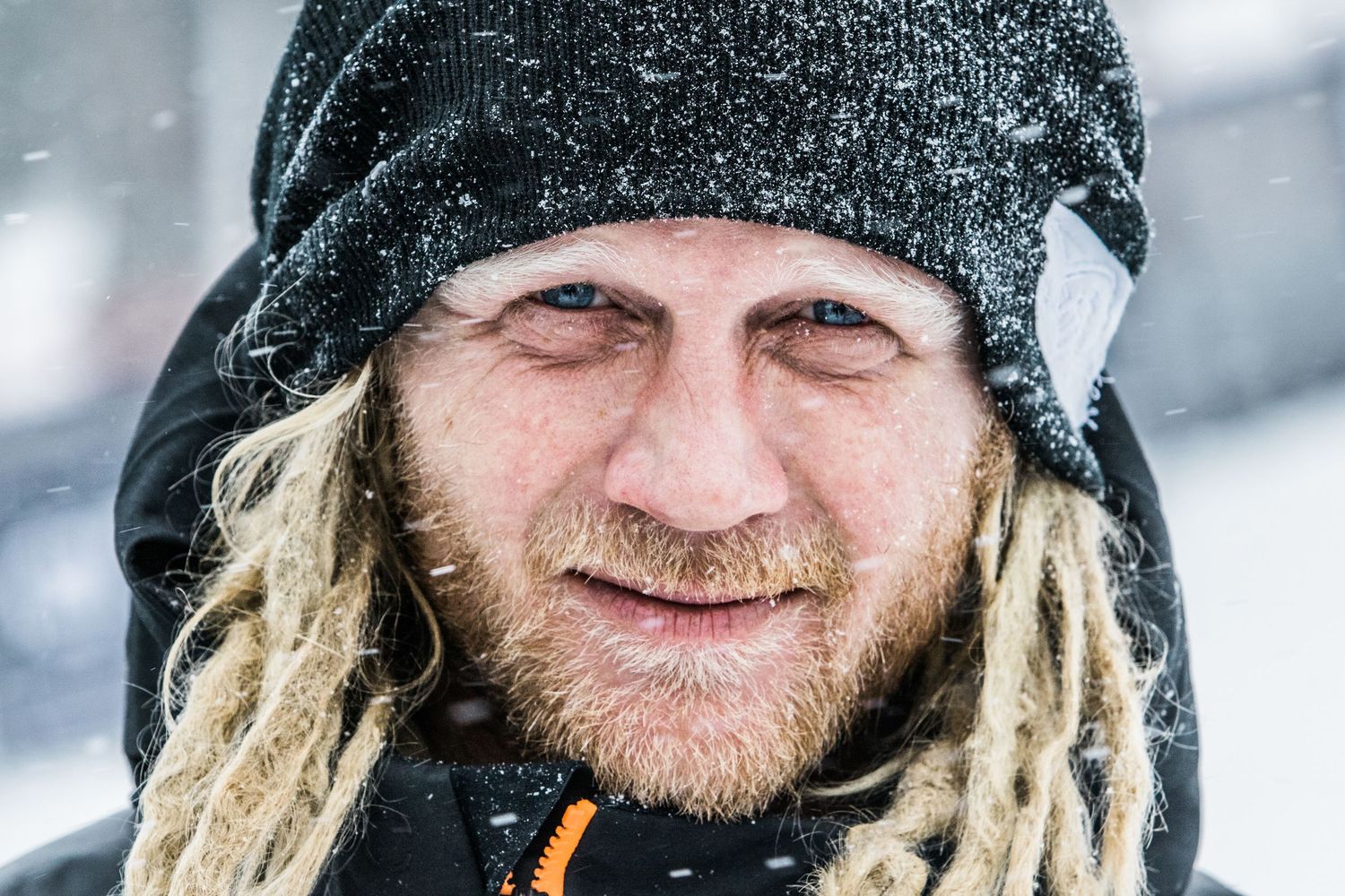 A person wearing a black beanie cap smiles while looking directly at the camera in soft lighting.