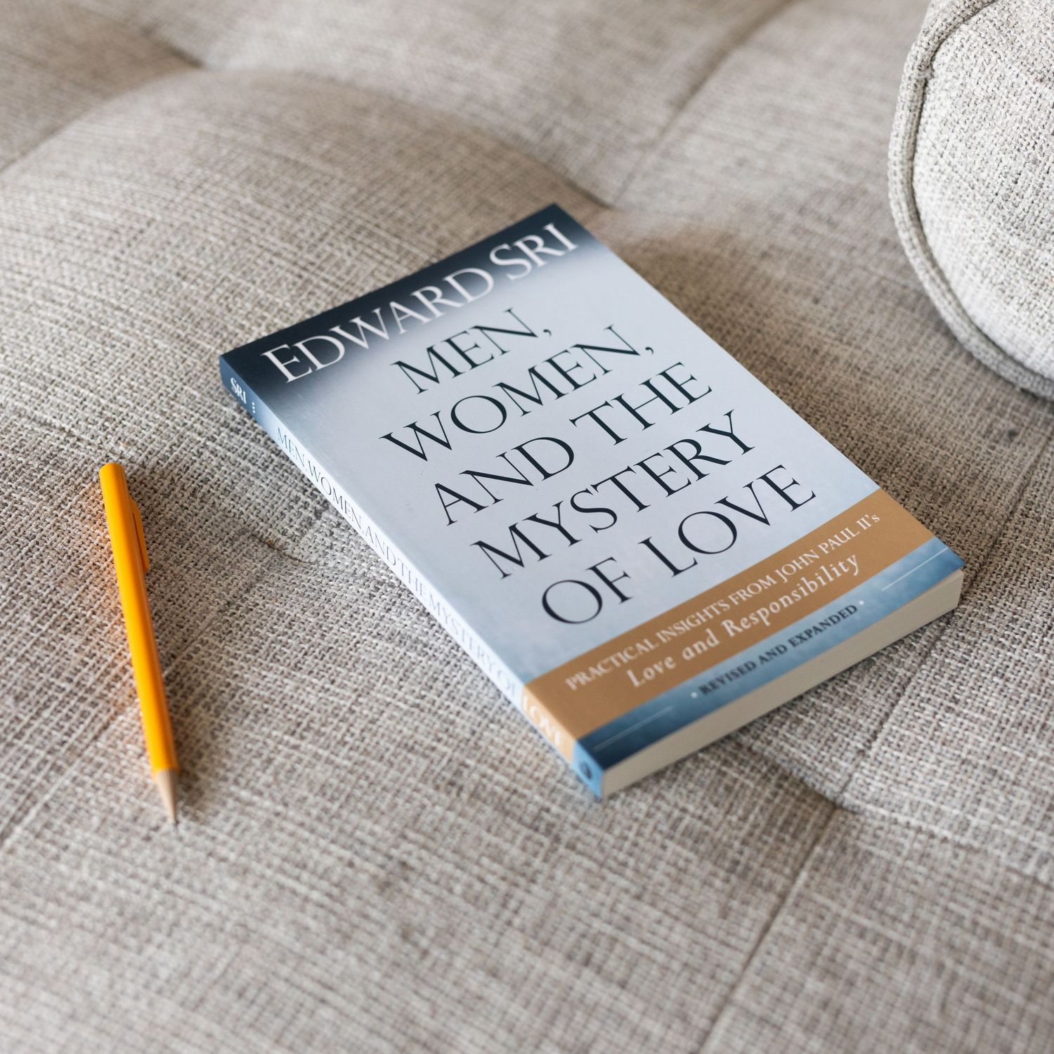 Book 'Men, Women, and the Mystery of Love' by Edward Sri resting on a beige couch beside a yellow pencil.
