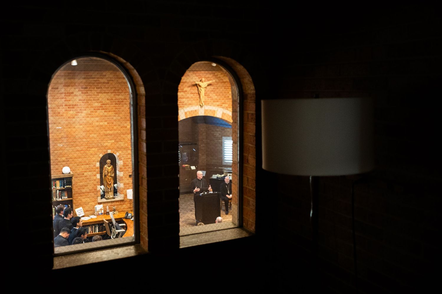View through arched windows into a warmly lit brick chapel with robed figures and a cross on the wall.