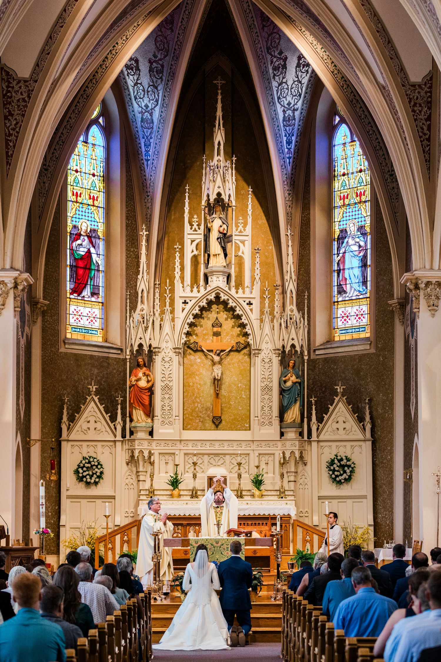 Bride and groom kneel at ornate Catholic church altar with stained glass windows during wedding ceremony.