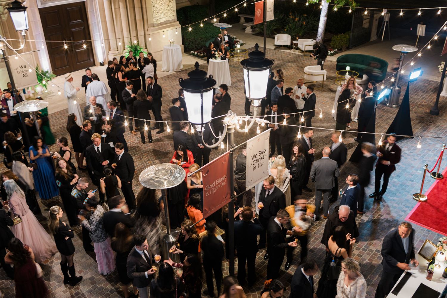 Crowded social event with string lights overhead showing people mingling and networking in an indoor venue.