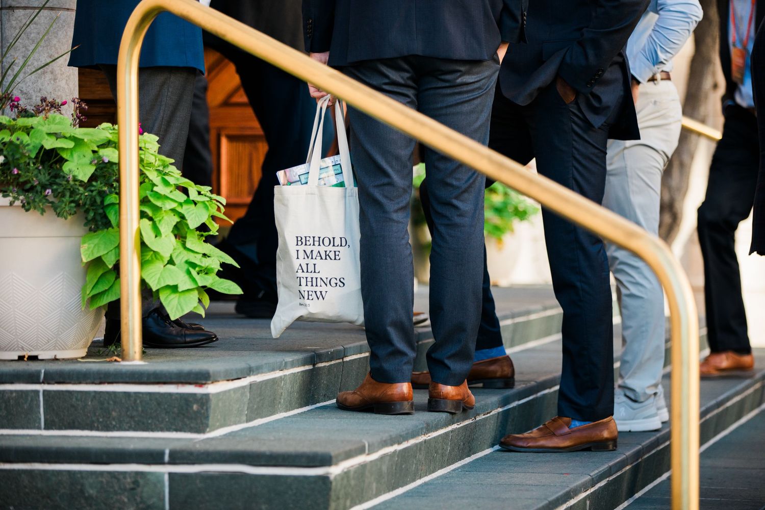People standing on steps with a tote bag reading 'Behold I Make All Things New' near a railing.