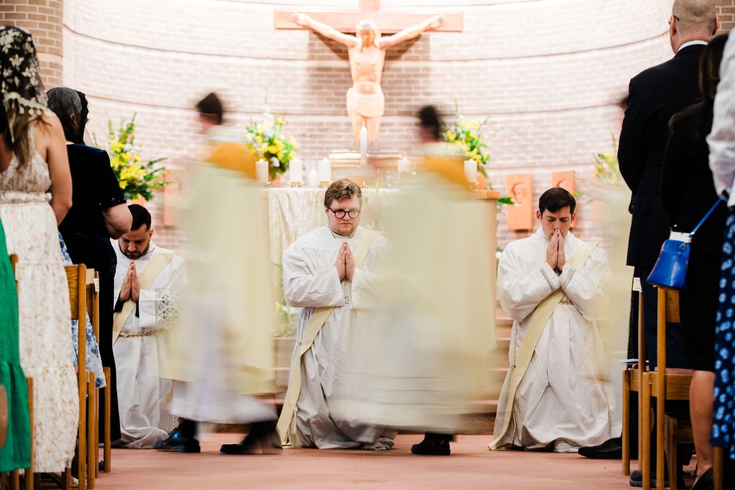 Clergy kneeling in prayer during a Catholic Mass ceremony inside a church with a crucifix altar.