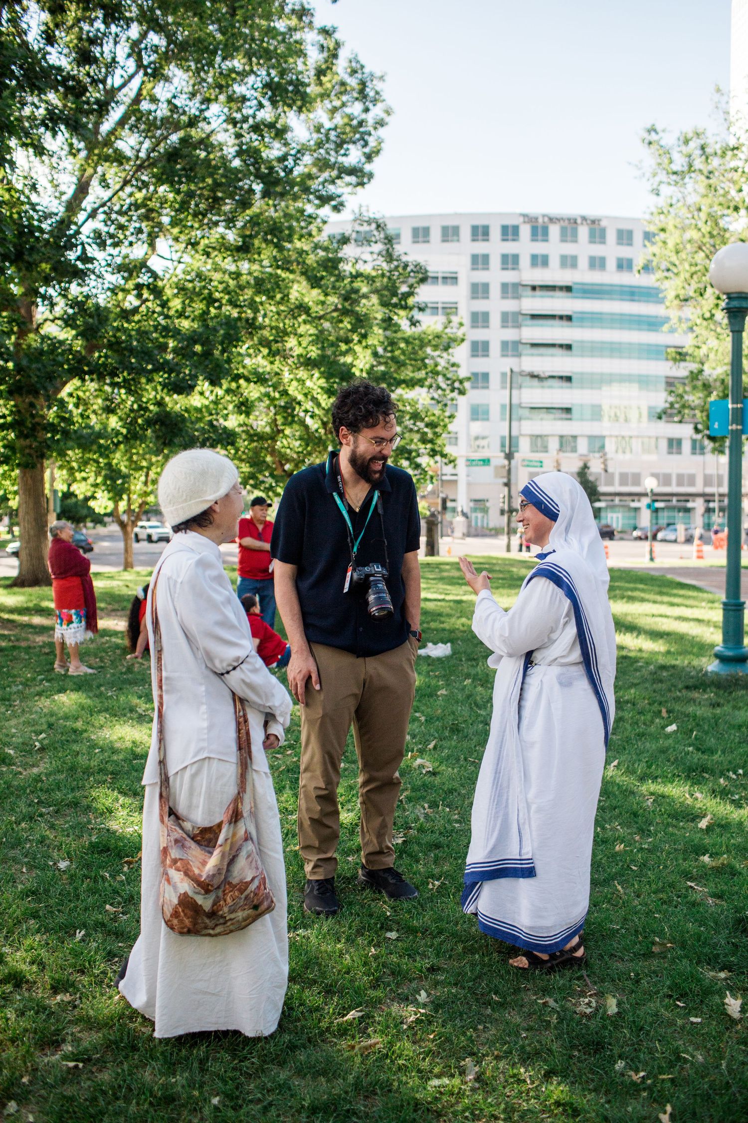 A nun in white habit converses with a man and woman in a sunny park near a modern building.
