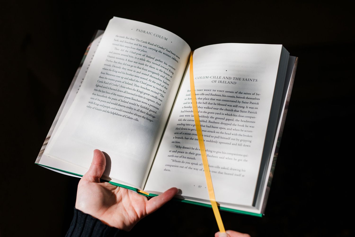 Hands holding an open book with a yellow pencil bookmark against a dark background.