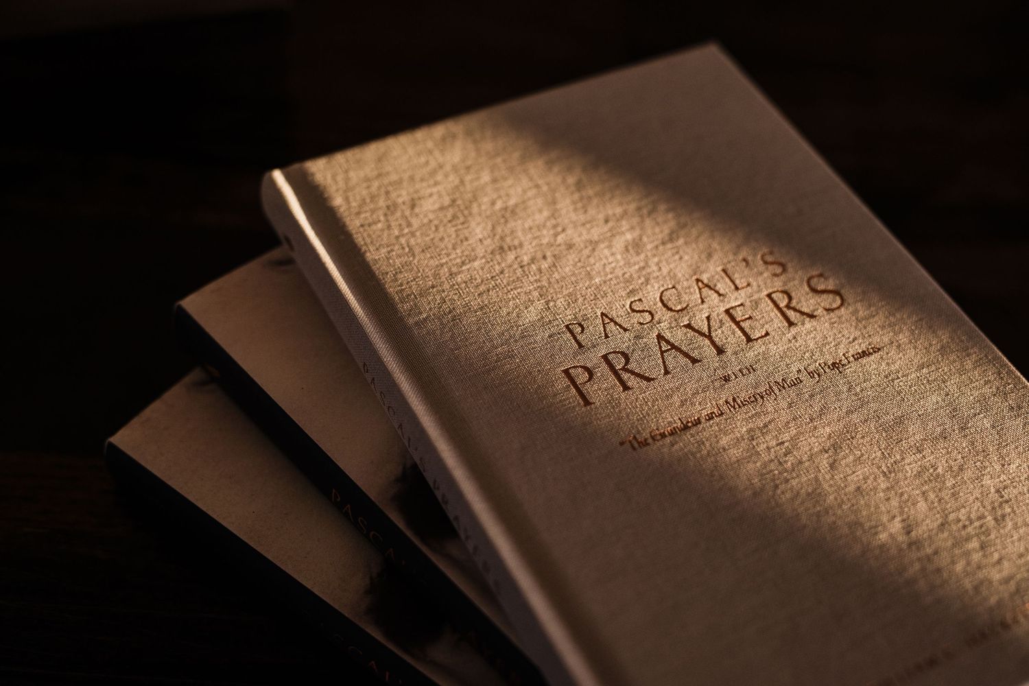 Stack of Pascal's Prayers books with elegant gold embossed text on textured cover in dramatic lighting.