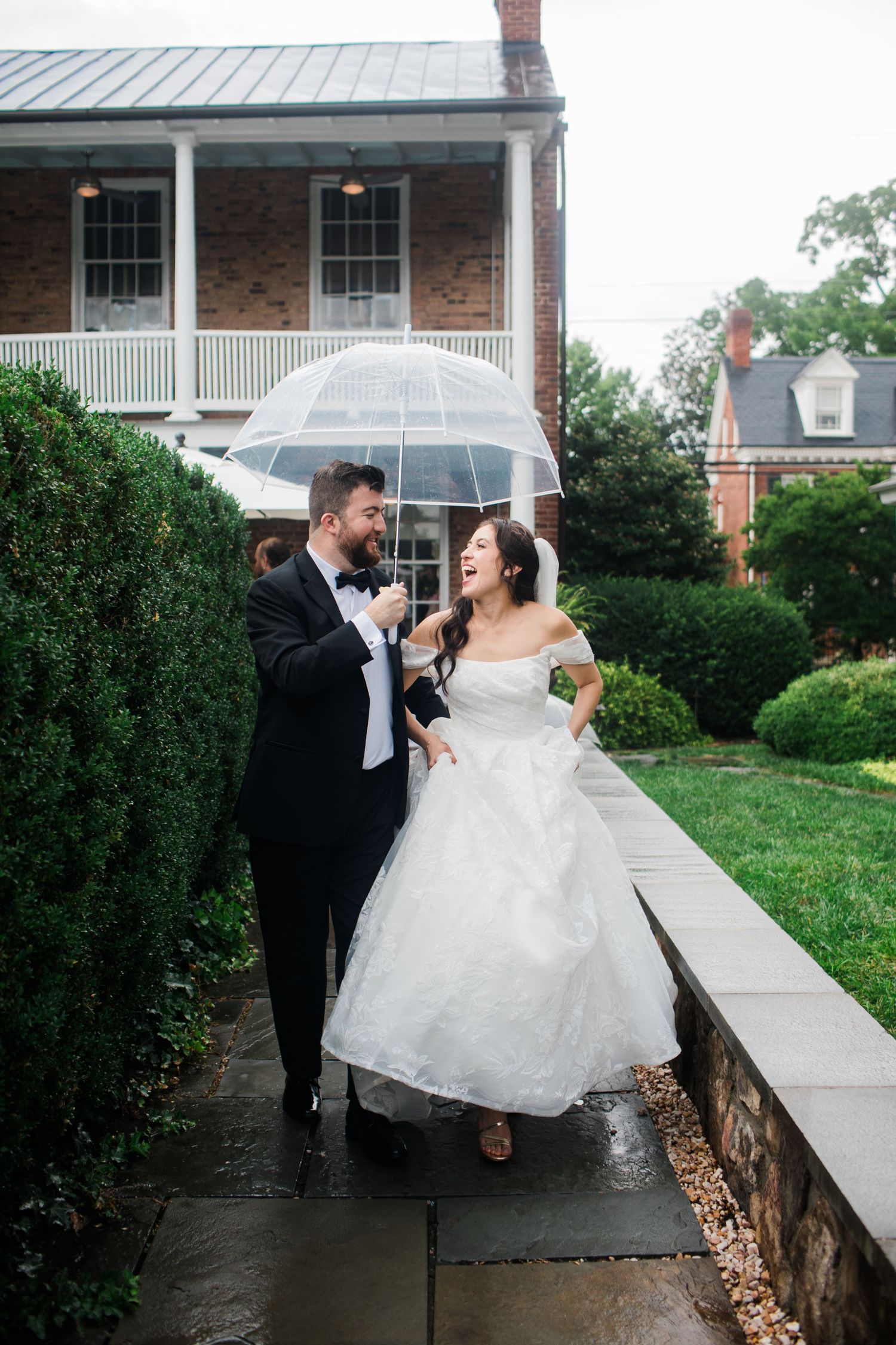 Bride and groom laughing under clear umbrella on rainy wedding day outside brick venue.