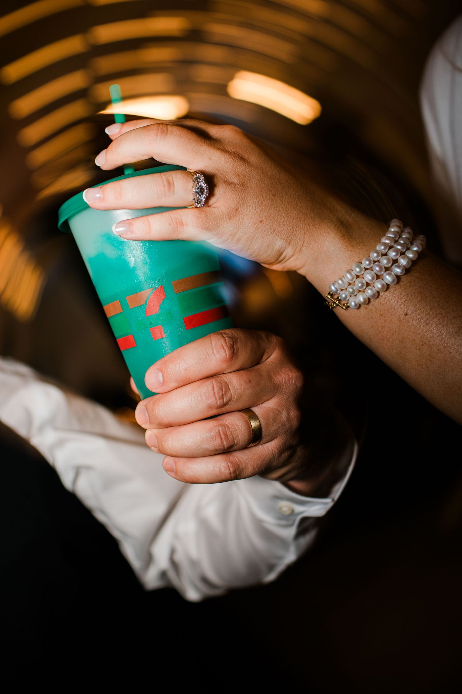 Close-up of bride and groom hands holding teal cup, wearing wedding rings and pearl bracelet.