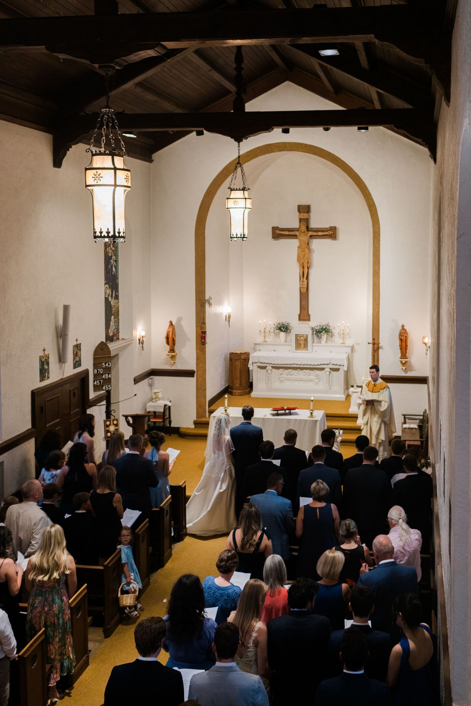 A wedding ceremony inside a small Catholic chapel with guests seated in pews as the bride and groom stand at the altar.