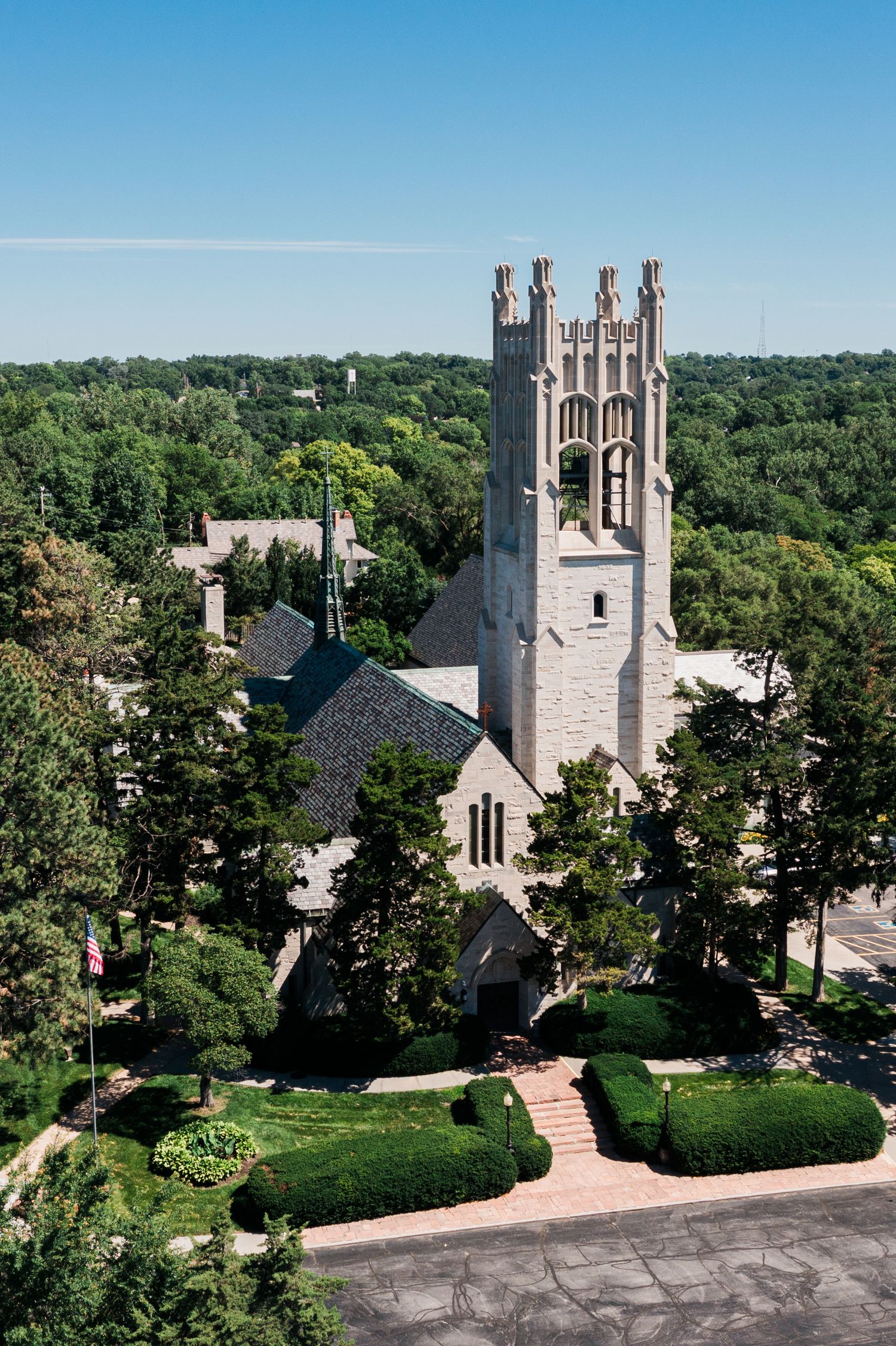Aerial view of a historic stone church with Gothic tower surrounded by lush green trees under a clear blue sky.
