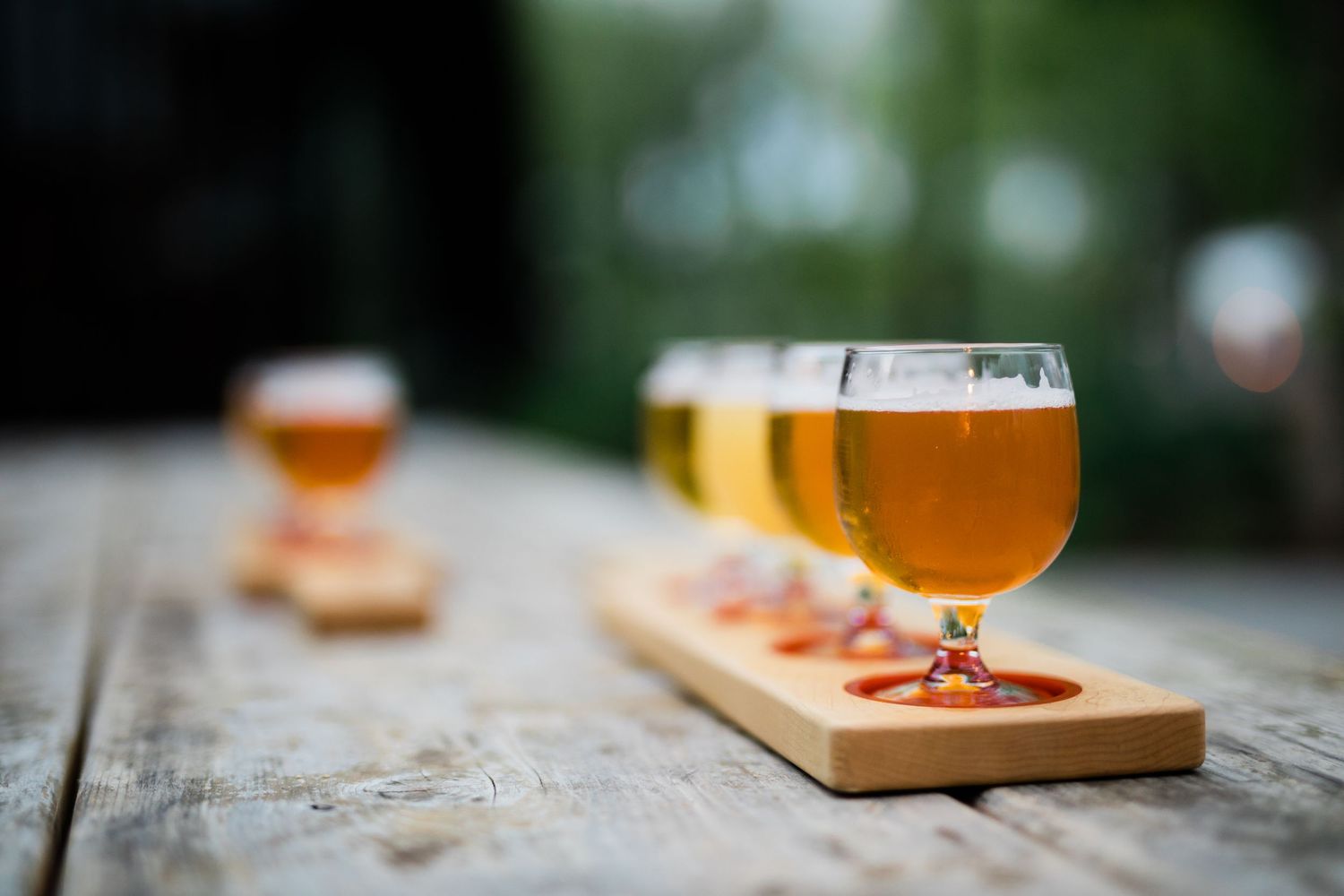Craft beer glasses with orange-hued beer sit on a wooden outdoor table with blurred background.